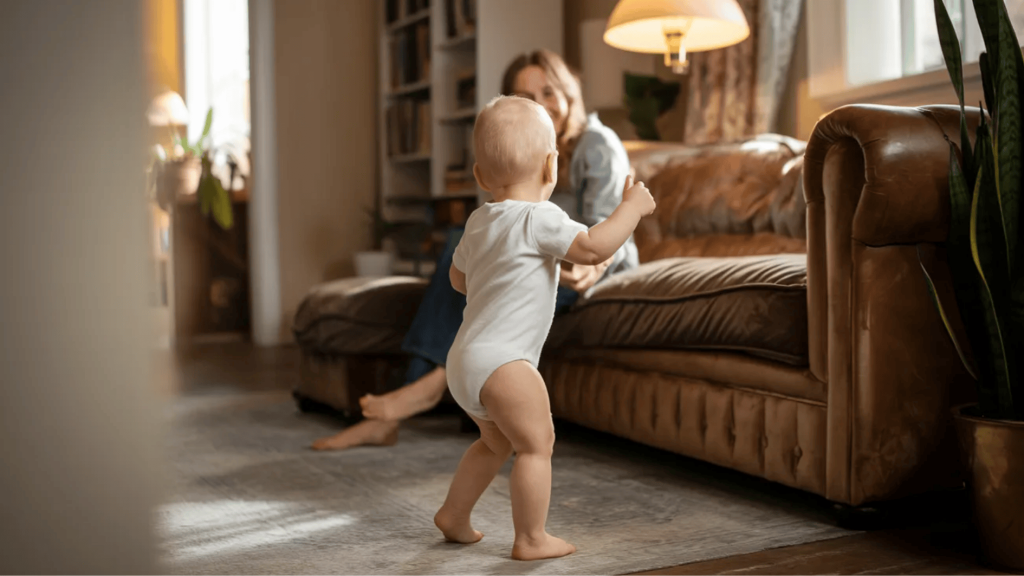 baby taking first steps toward parent on couch in a cozy living room with warm lighting