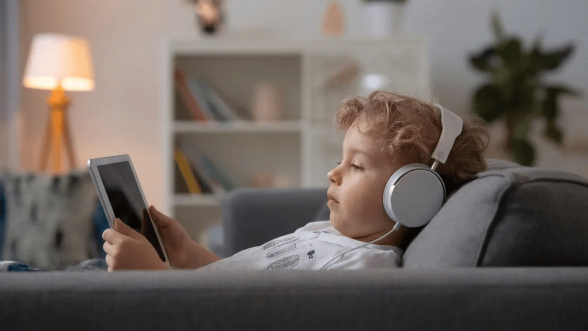 child lying on a sofa wearing headphones and listening to an audiobook on a tablet in a cozy living room
