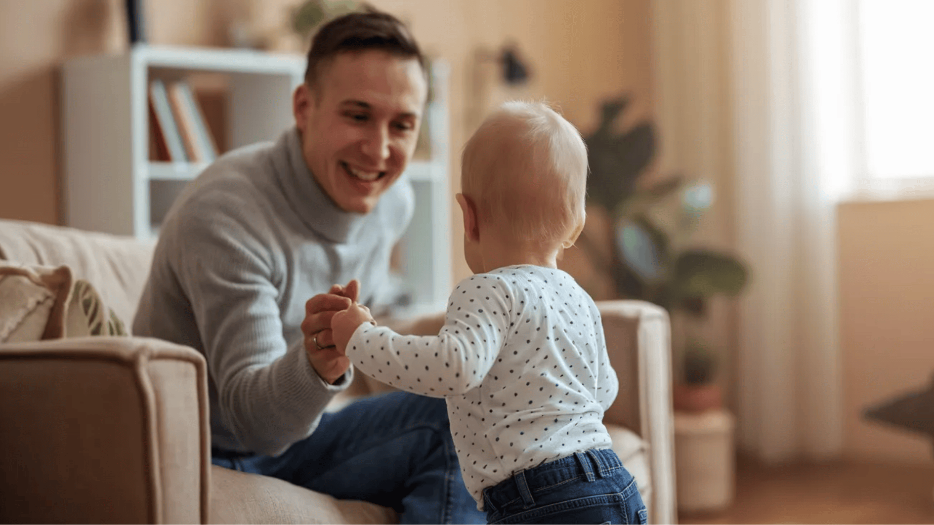 father smiling and holding baby’s hands as the child stands in a warm, cozy living room