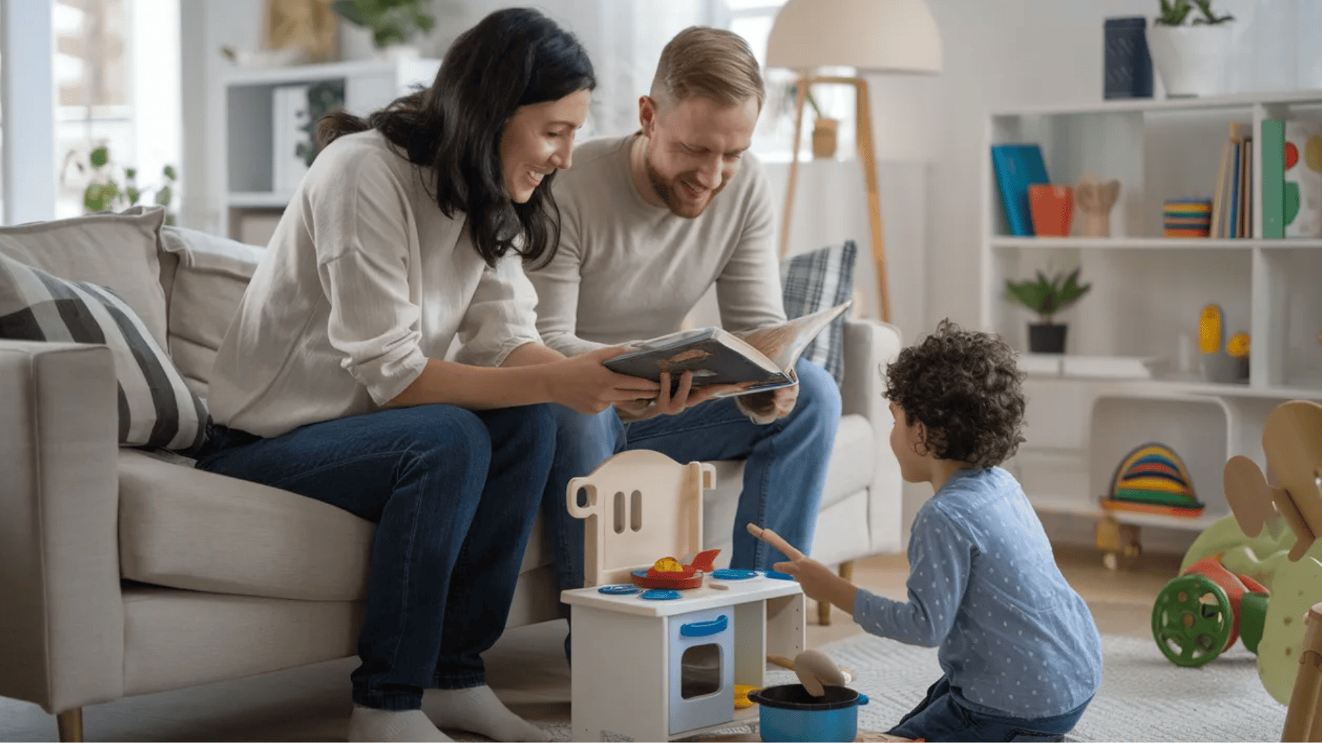 parents reading a picture book on sofa while young child plays with toy kitchen set in cozy living room