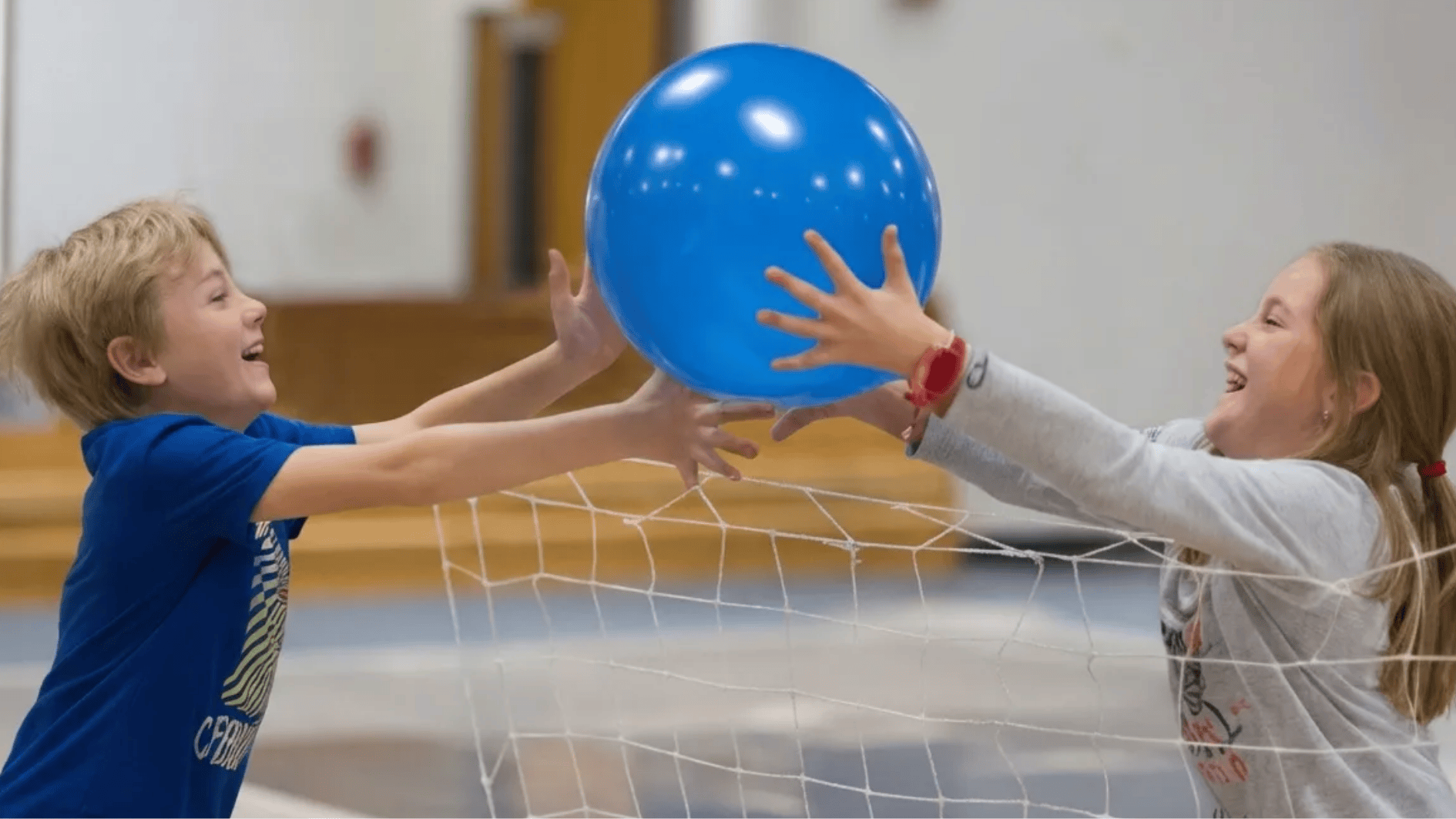 two kids laughing and reaching for a blue ball over a net while playing an indoor game together