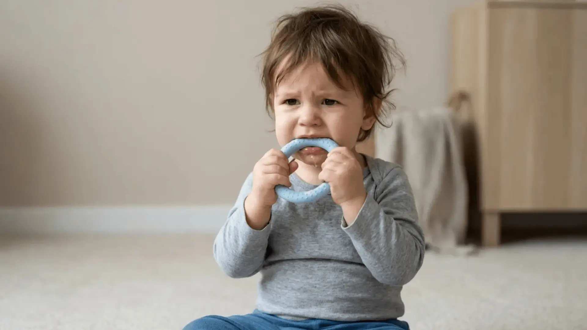 Toddler sitting and chewing on an object with slightly fussy expression indoors