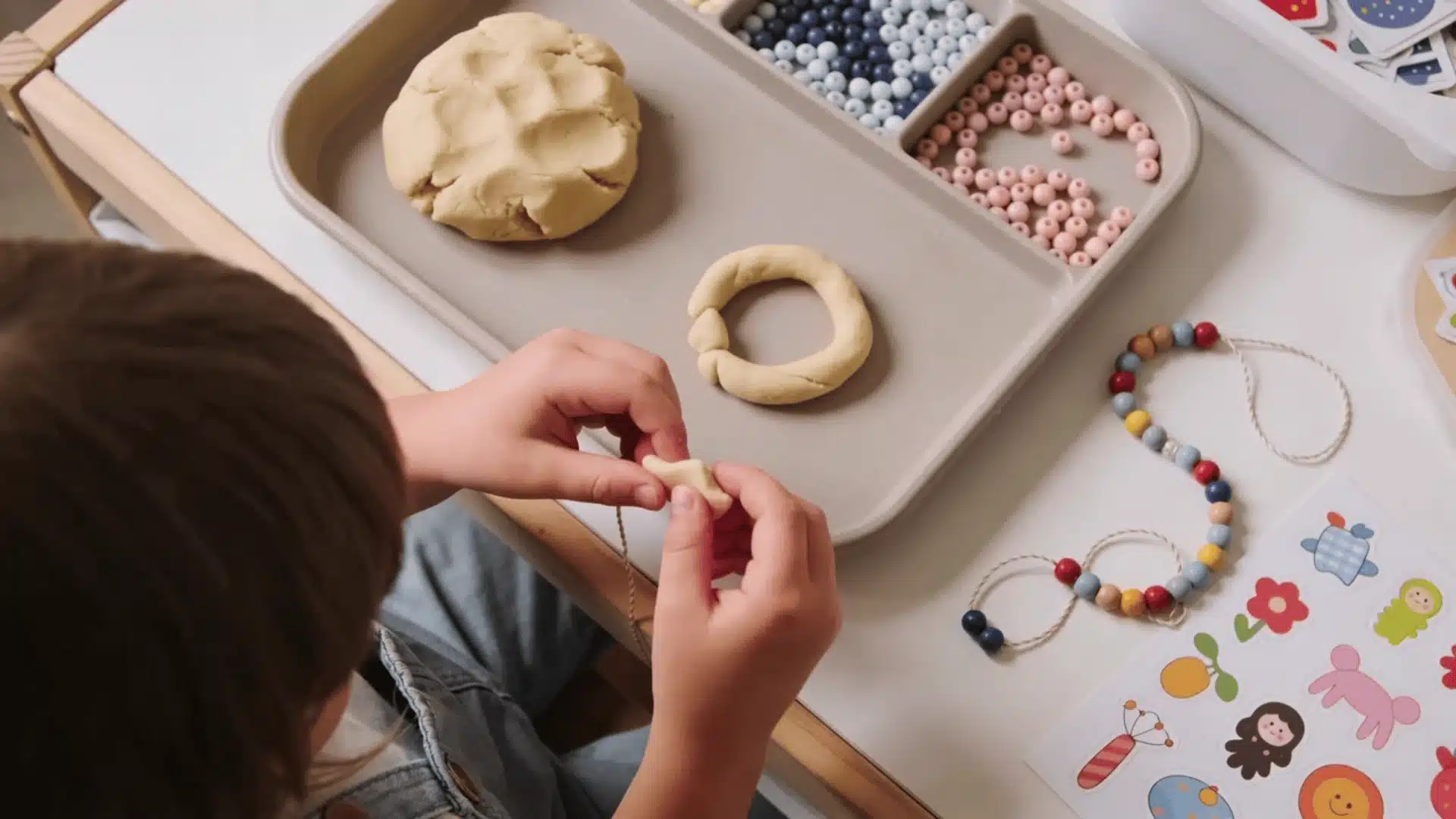 Toddler using playdough, beads, and stickers on a tray for fine motor play
