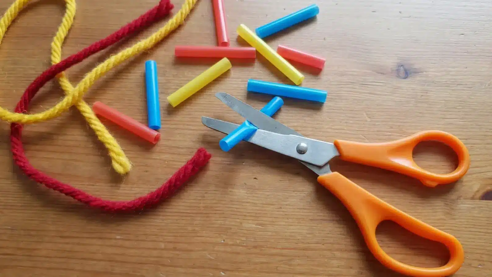 Child-safe scissors cutting small pieces of straws and yarn on a table