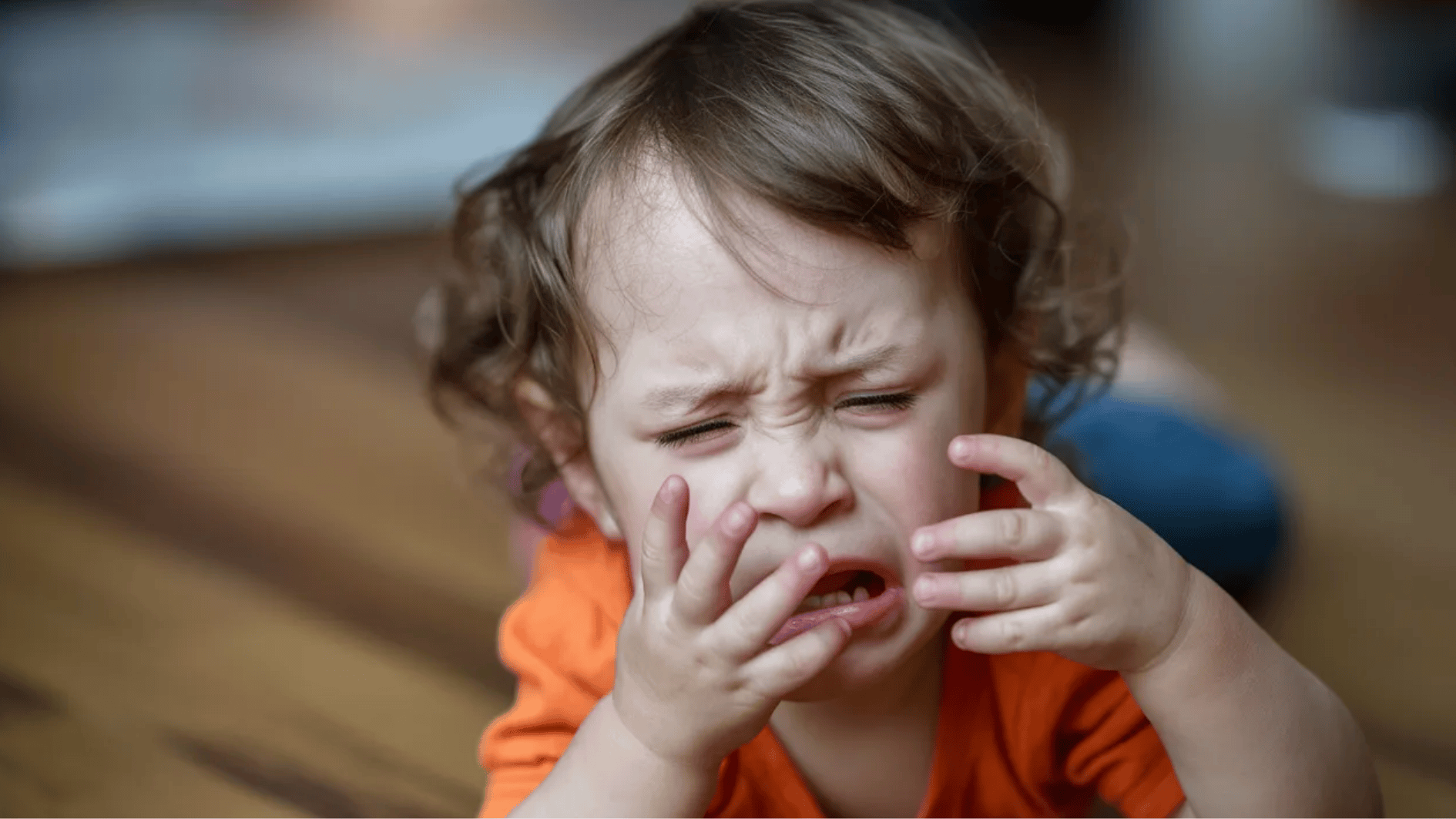 close-up of crying toddler with hands on face, expressing distress in a softly blurred indoor setting
