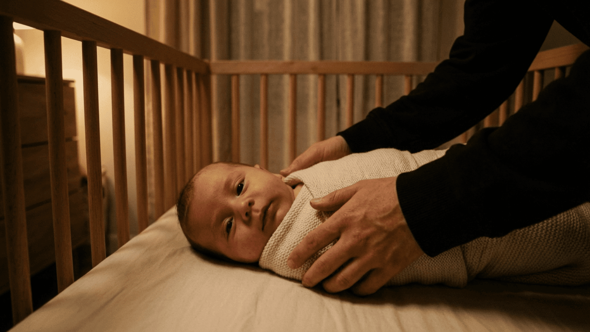 full baby placed into crib by parent hands in same warm dim bedroom with calm expression and eyes slightly open