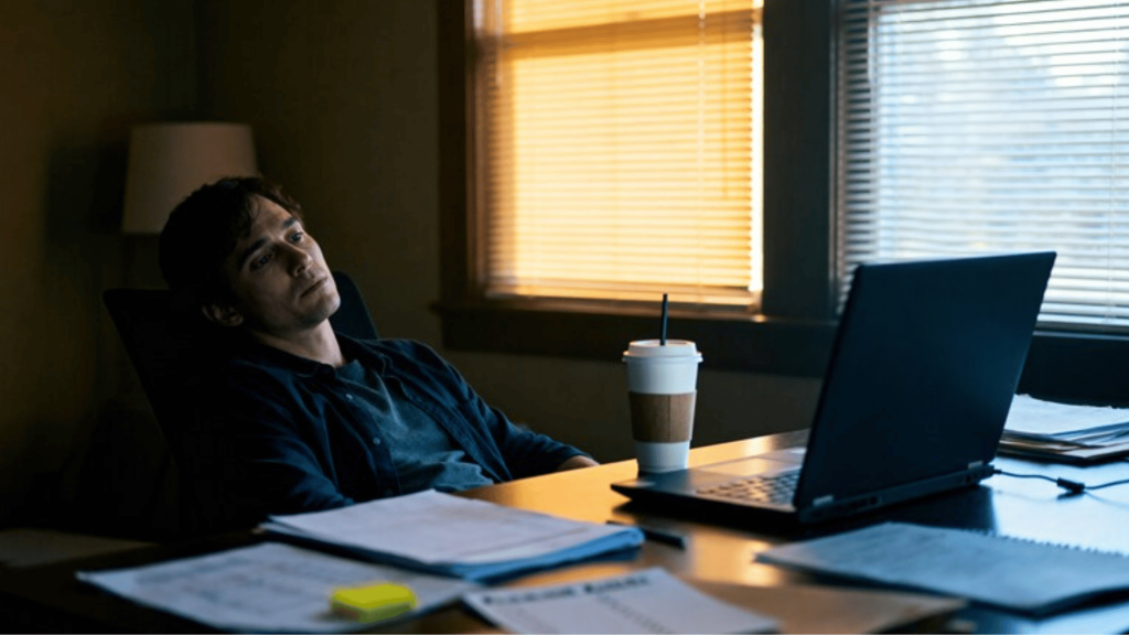 tired man sitting at desk with laptop and paperwork, showing work stress, fatigue, and burnout in a dimly lit home office