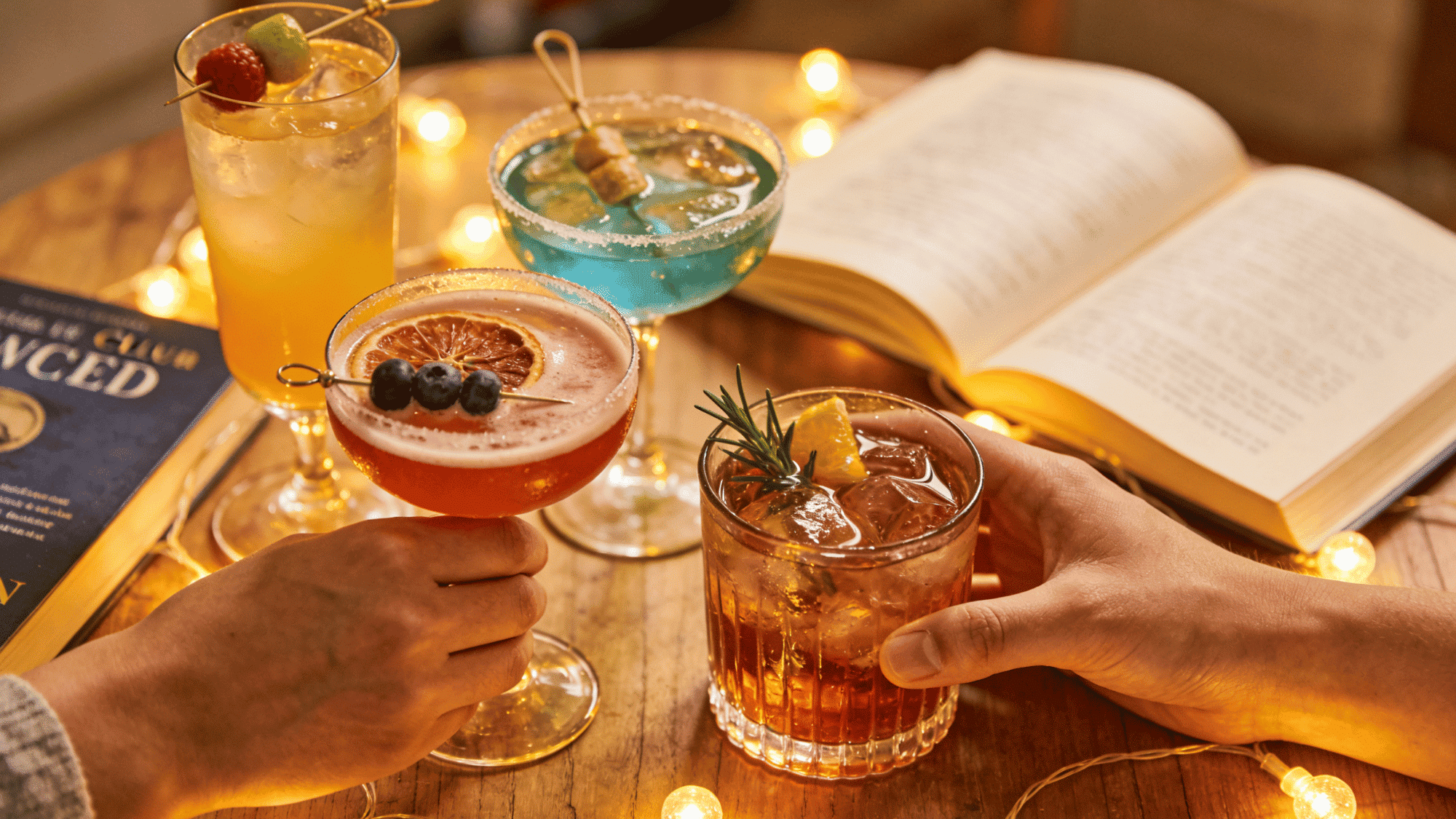 cozy book club mocktail station with colorful drinks, recipe cards, an open book, and hands reaching for glasses in warm light