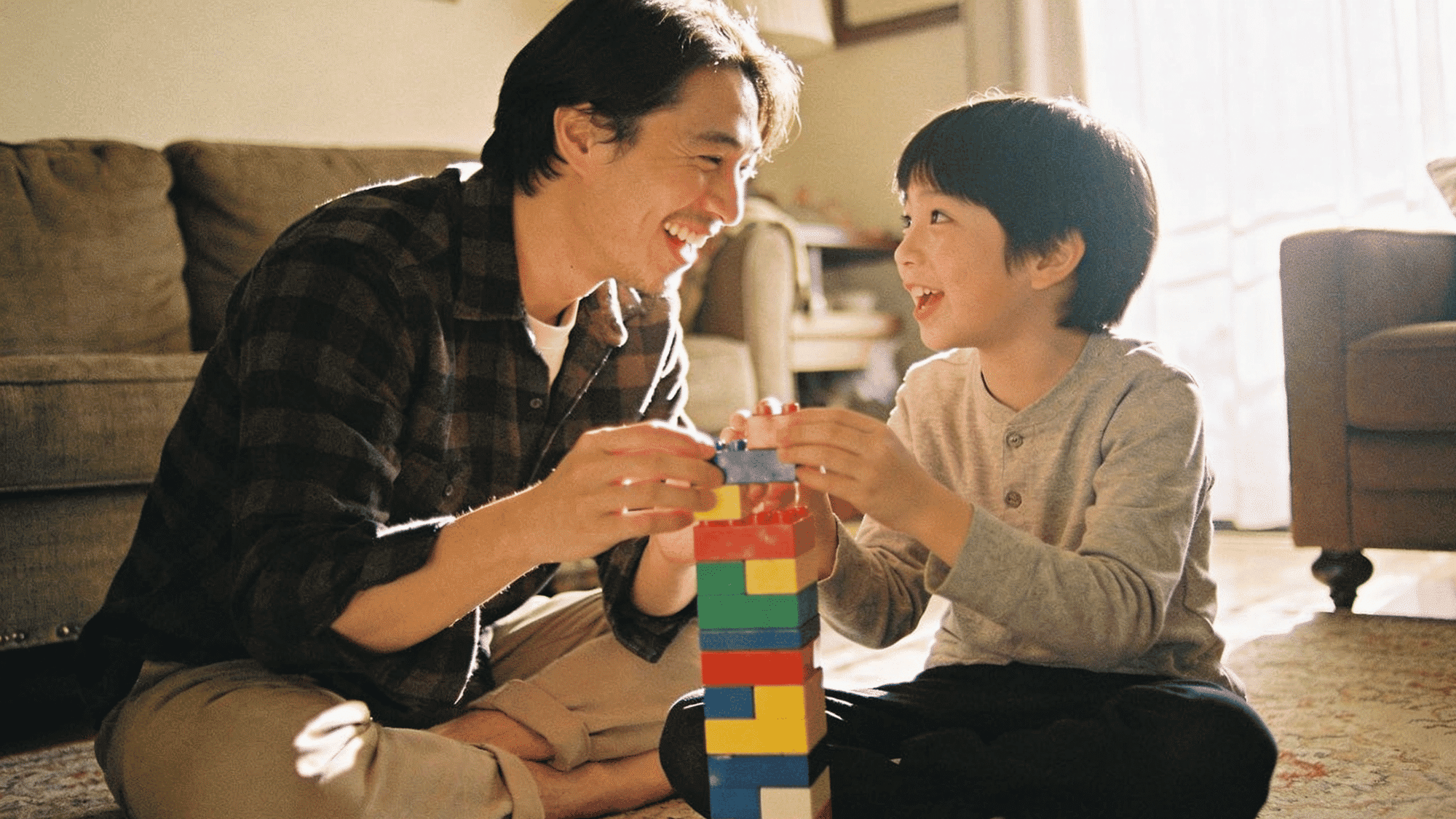 father and young son smiling while building colorful block tower together on cozy living room floor at home