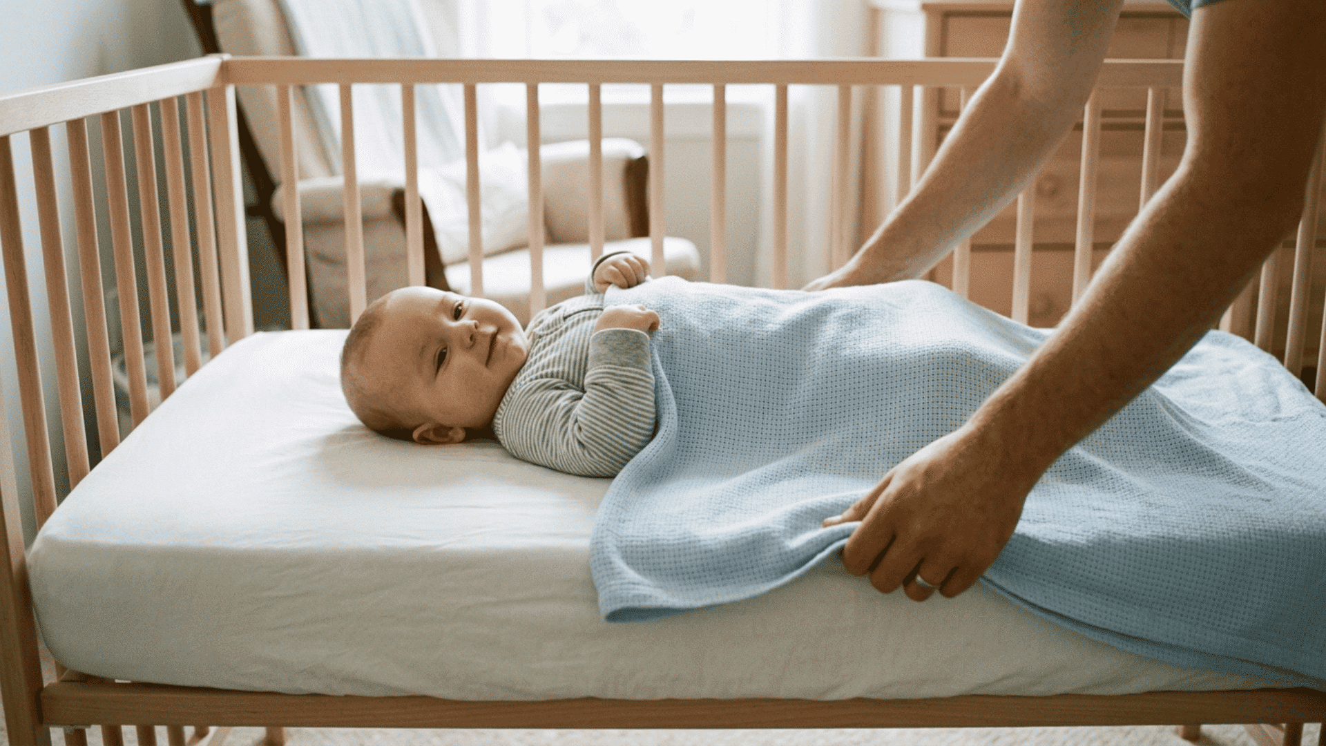 infant lying in crib with blanket below shoulders and parent hands tucking edges in a safe sleep setup