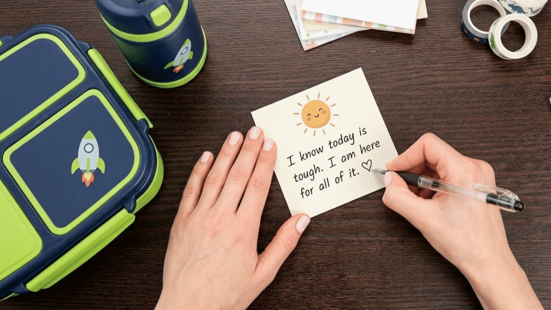 parent writing a sweet lunch box note beside colorful lunch containers on a wooden table