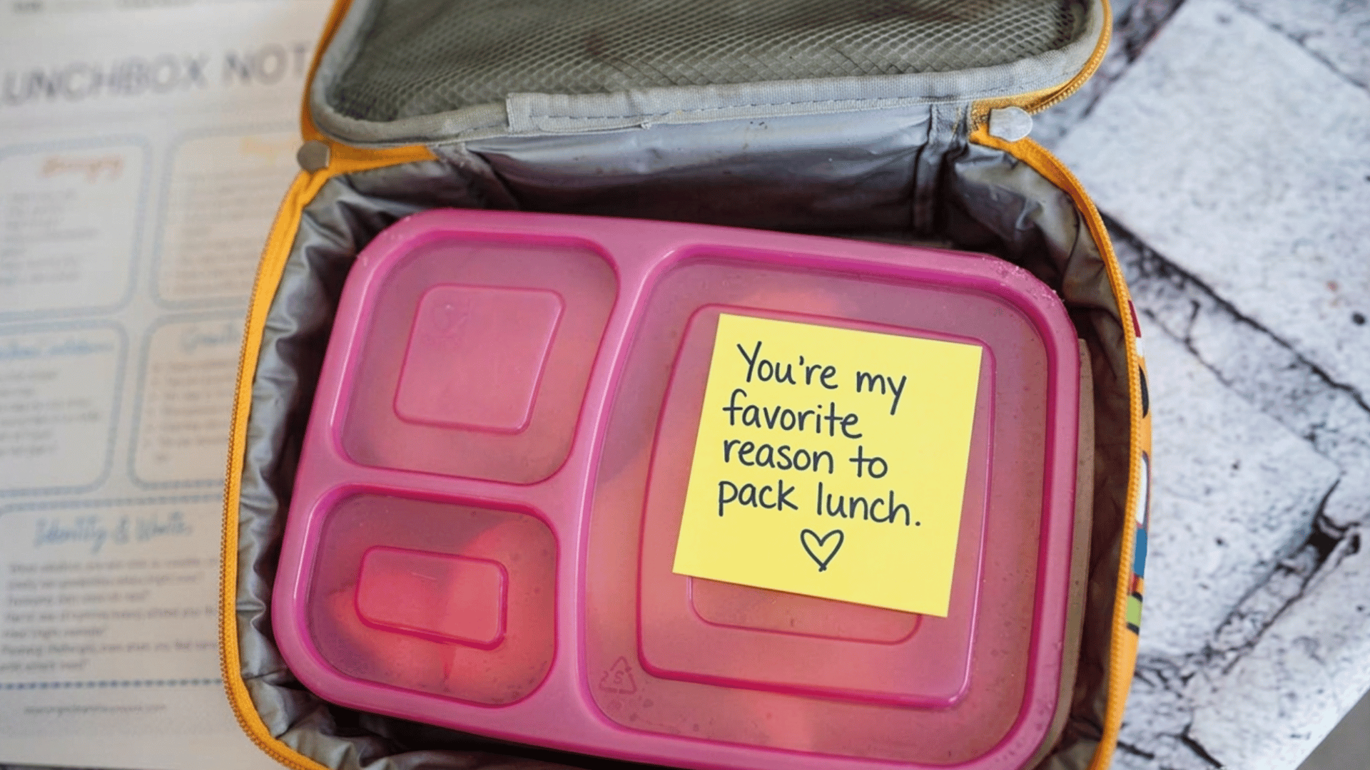 pink lunch box with colorful sticky note message inside, placed on a soft fabric background