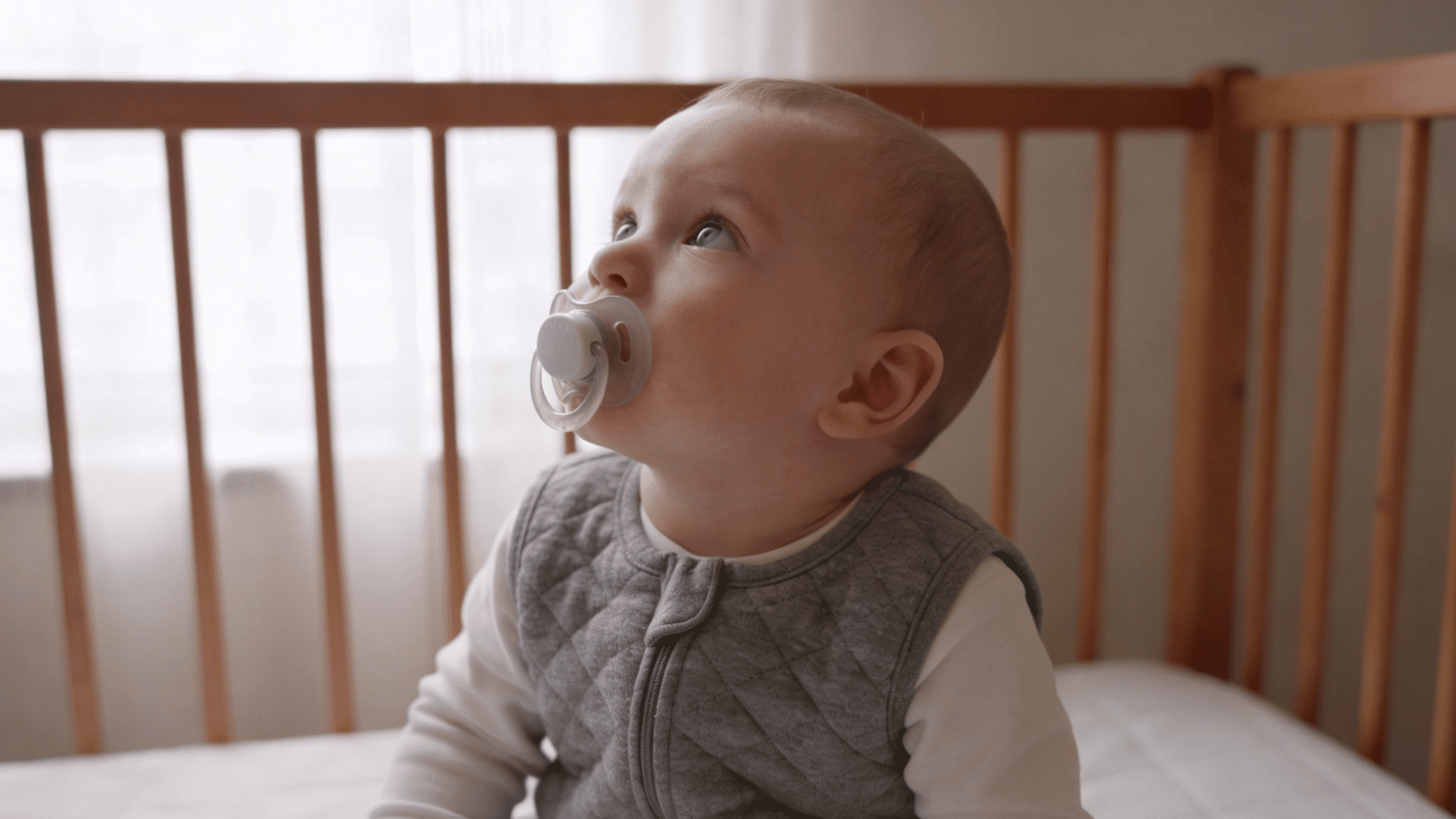baby with pacifier sitting in wooden crib, looking upward near soft window light in cozy nursery room