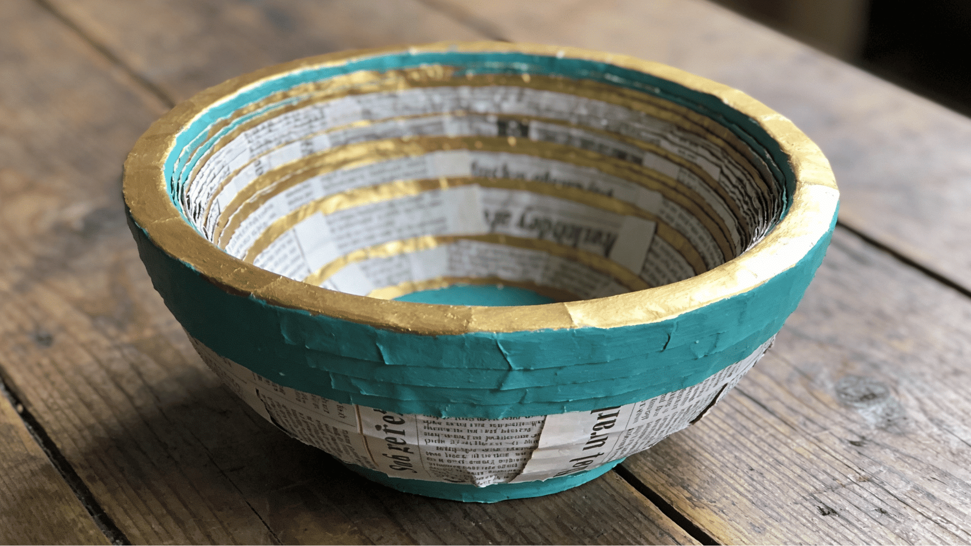 finished newspaper papier-mache bowl painted in teal and gold sitting on a rustic wooden table