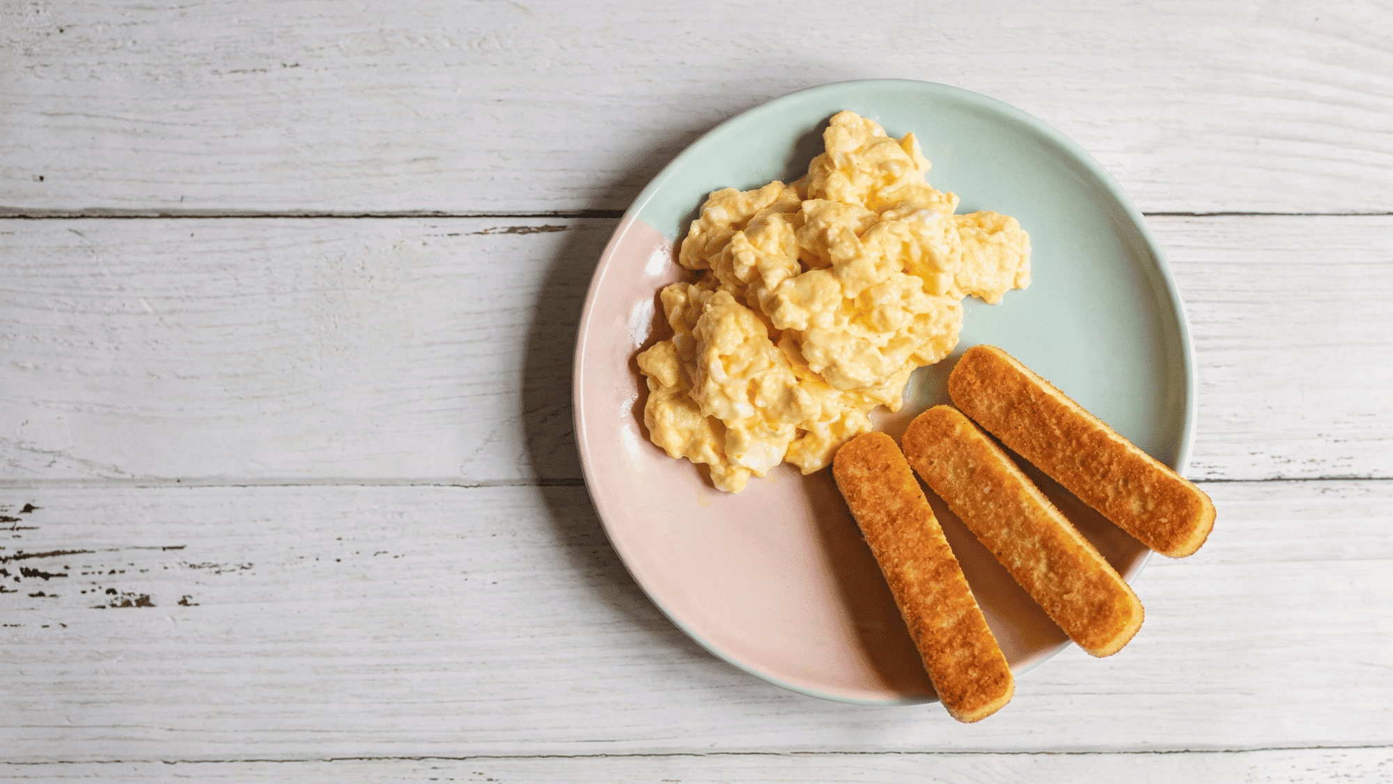 fluffy scrambled eggs in a white bowl beside golden toast sticks on a pastel blue plate