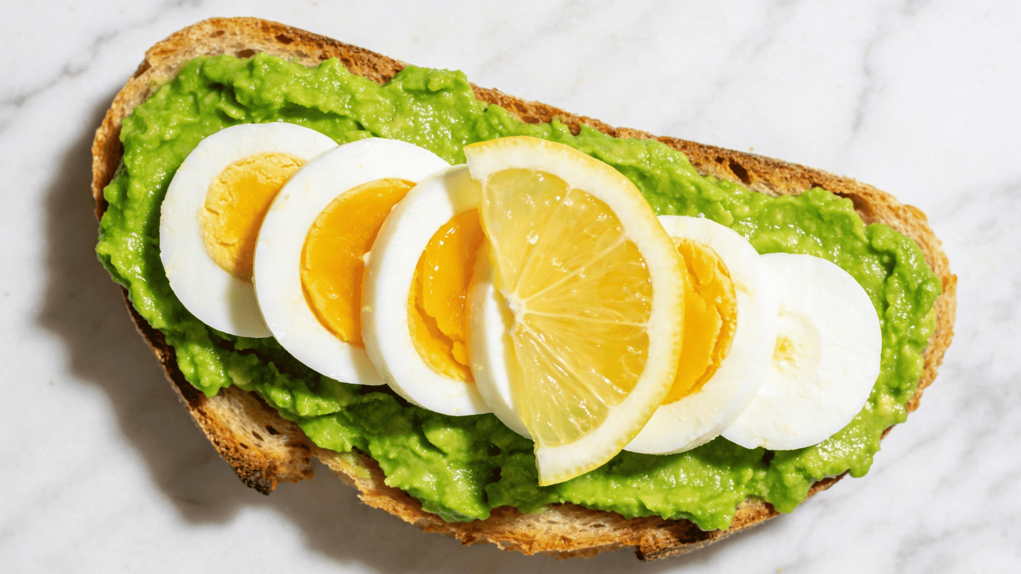 golden toast with mashed avocado, sliced boiled egg, lemon, and sea salt on a white plate