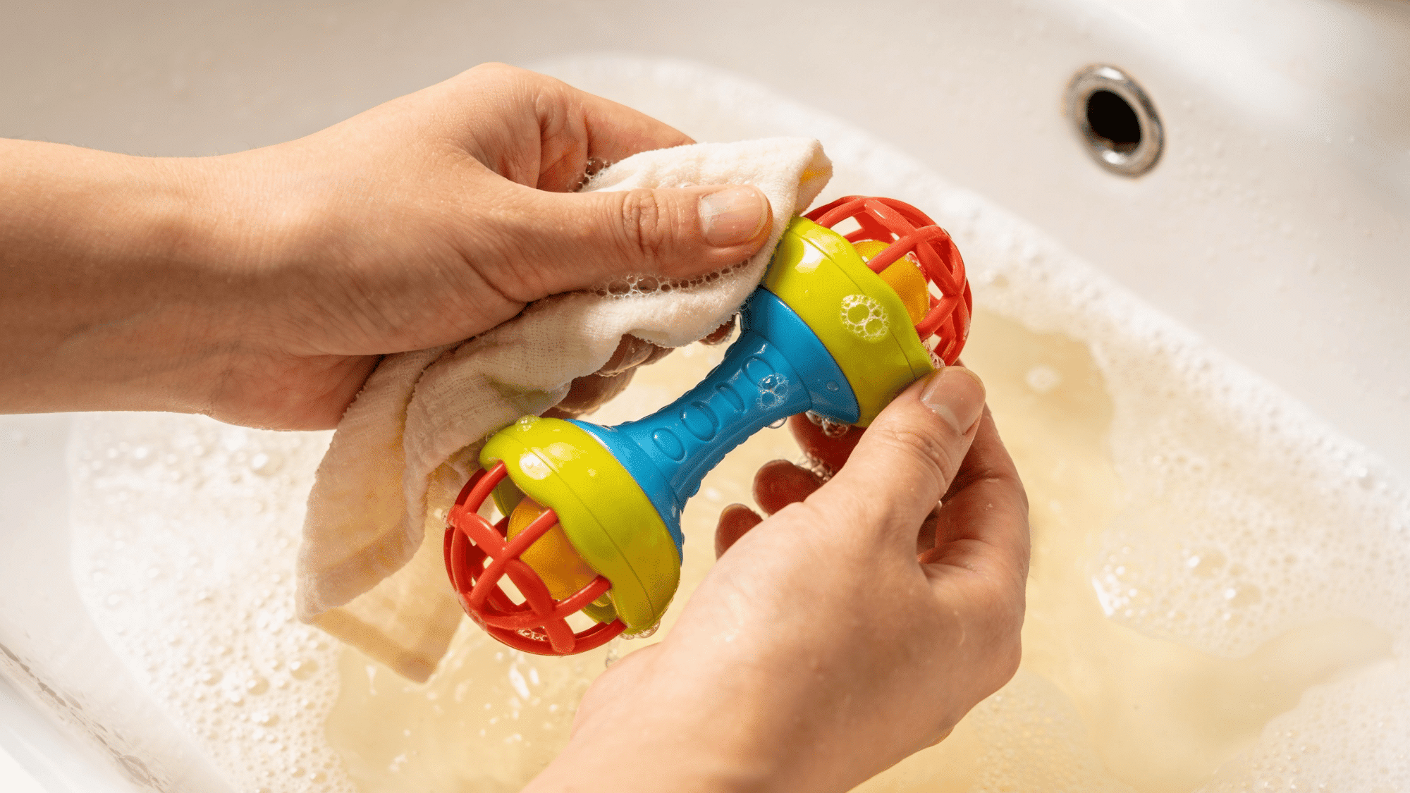 hand washing colorful baby rattle toy in warm soapy water in bathroom sink