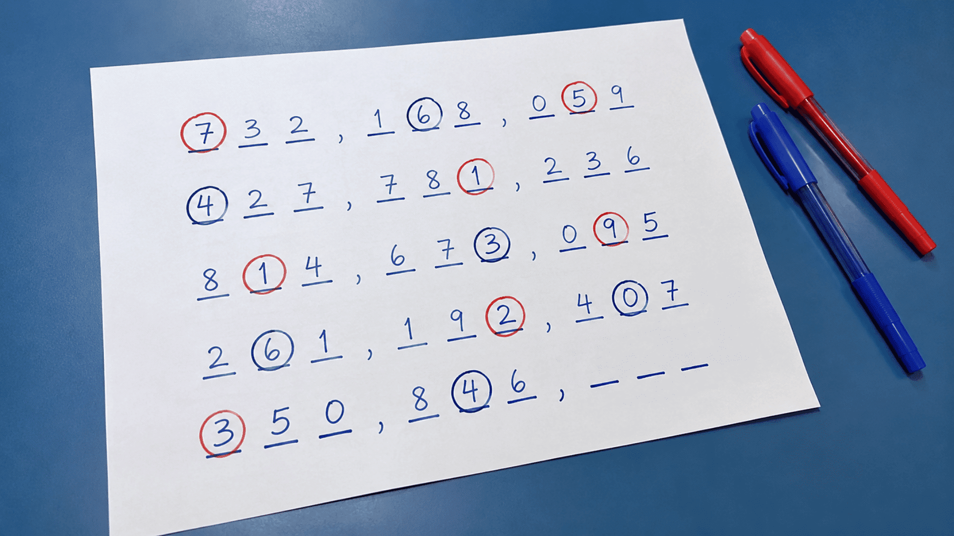 handwritten number puzzle on blue desk red and blue pens on side in a pen and paper game setup with half finished game on paper