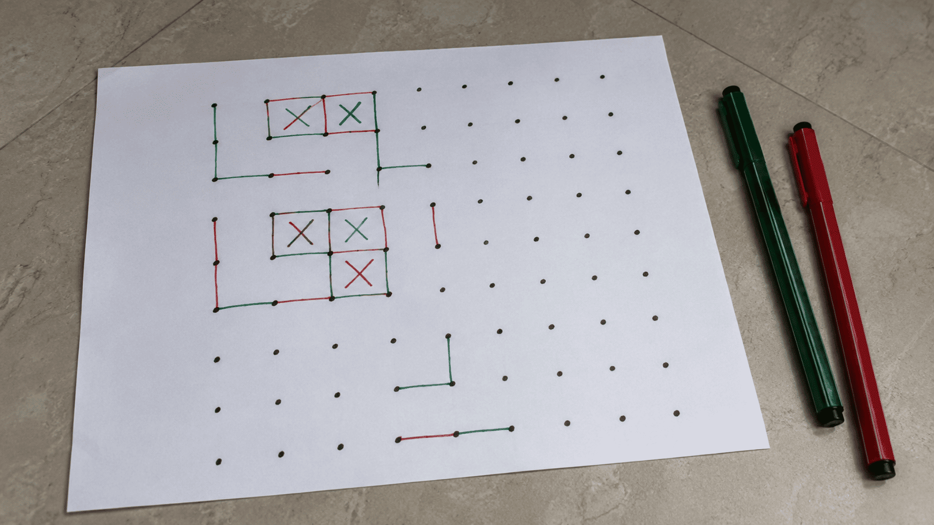 image of pen and paper game of dots and boxes game in pen and paper setup on a floor with green and red pen with grey floor visible