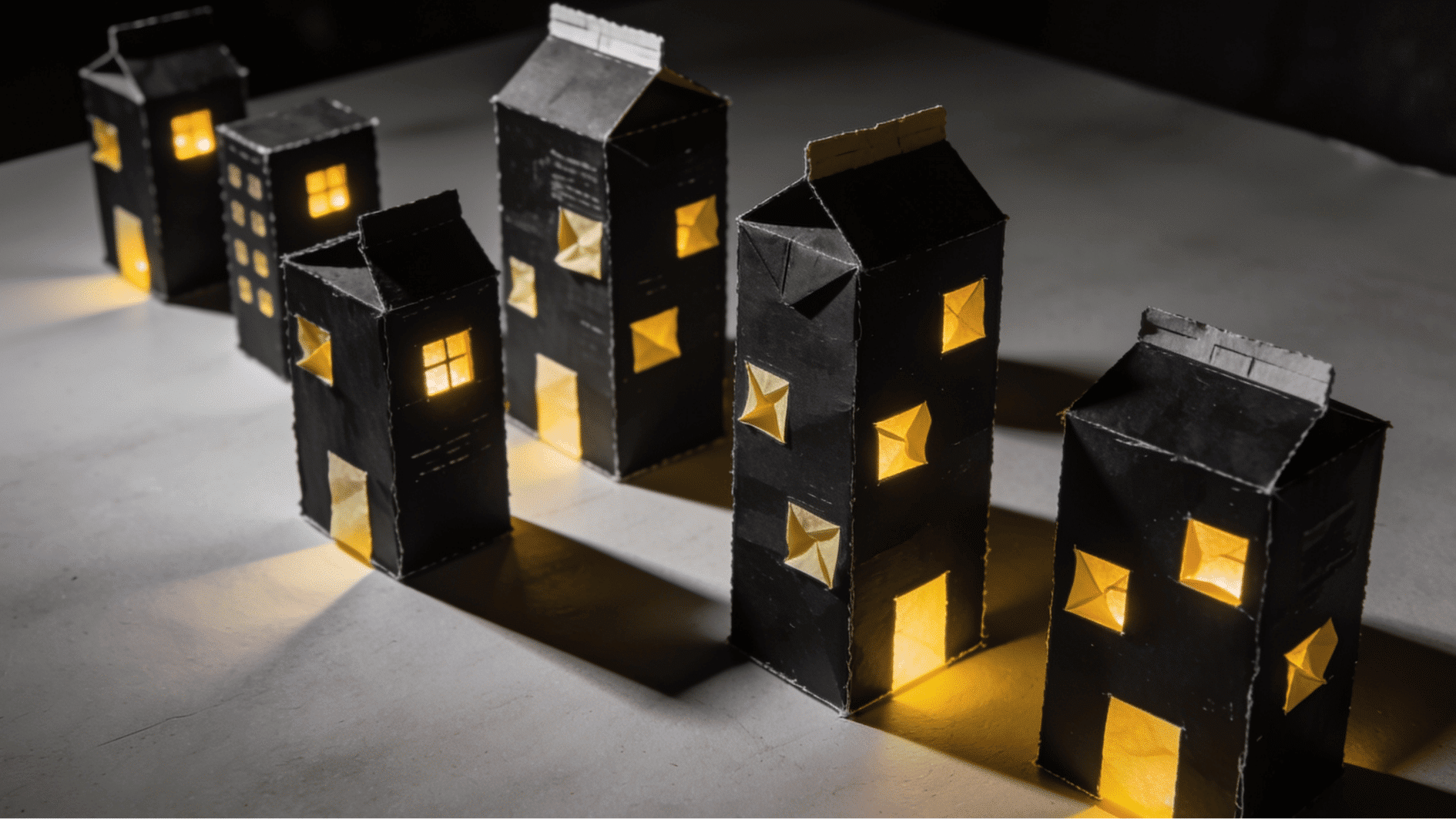 miniature cereal box cityscape painted black with glowing yellow tissue paper windows on a flat surface