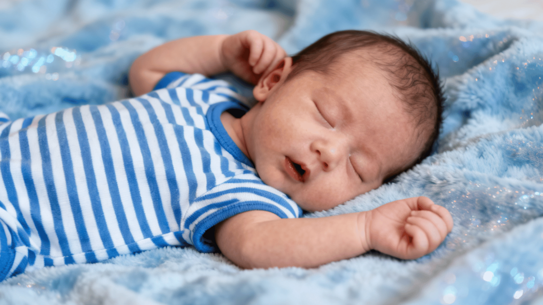 newborn baby sleeping on a fluffy blue blanket wearing a striped outfit with arms gently stretched and relaxed features