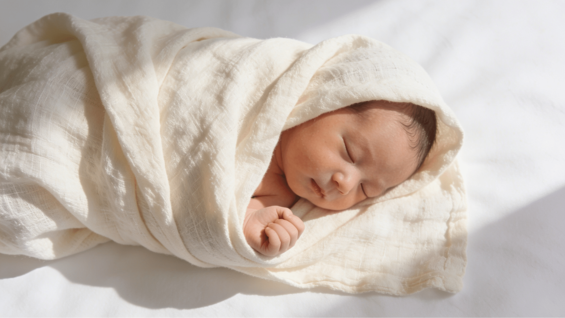 newborn baby sleeping peacefully swaddled in a soft cream muslin wrap on a white surface