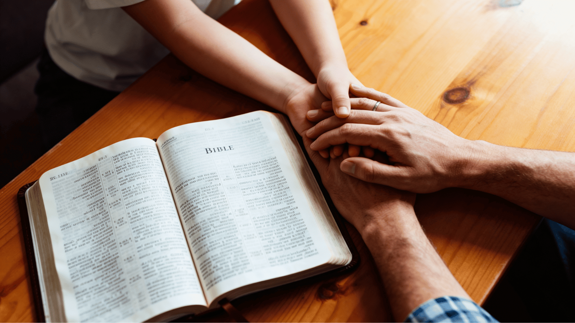 parent and child holding hands beside an open bible on a wooden table during prayer time