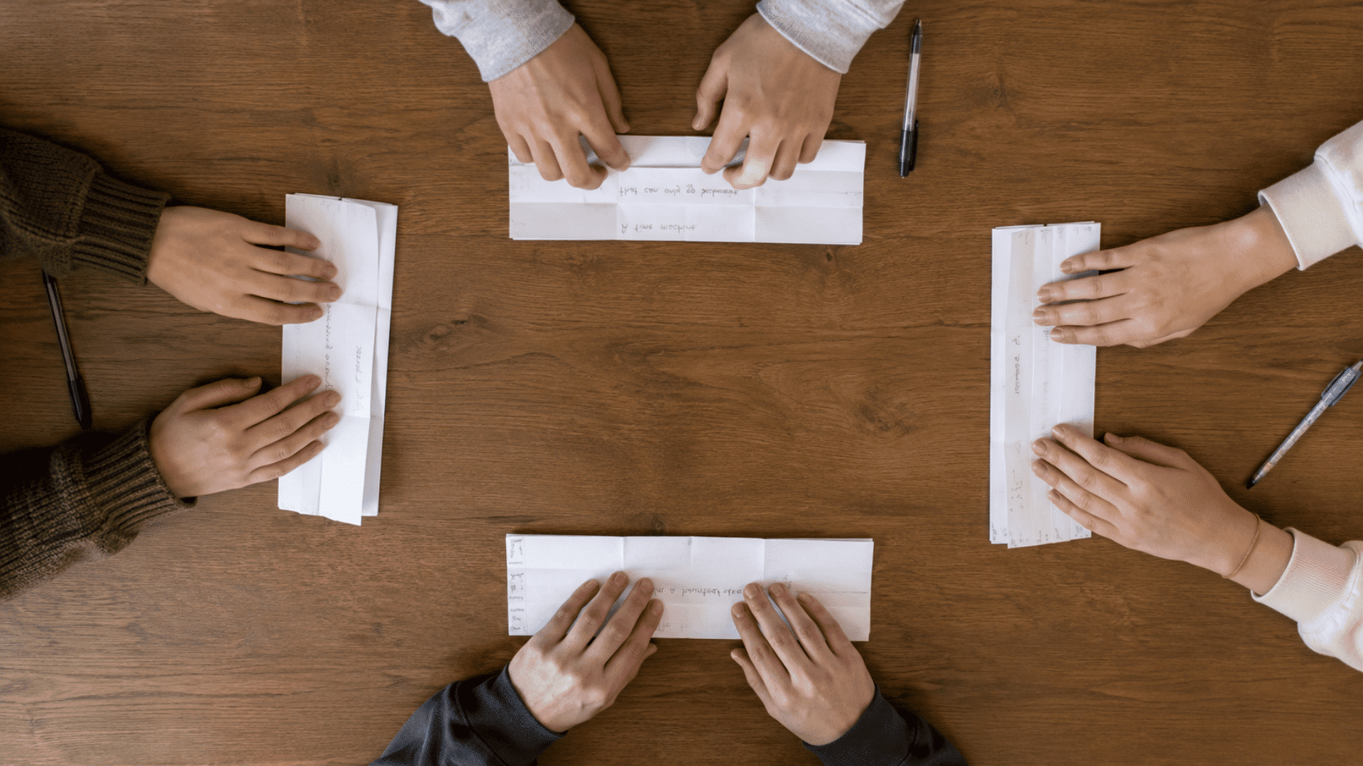 pen and paper group writing a game of consequences setup on a brown table with four pair of hands writing a story and folding their part