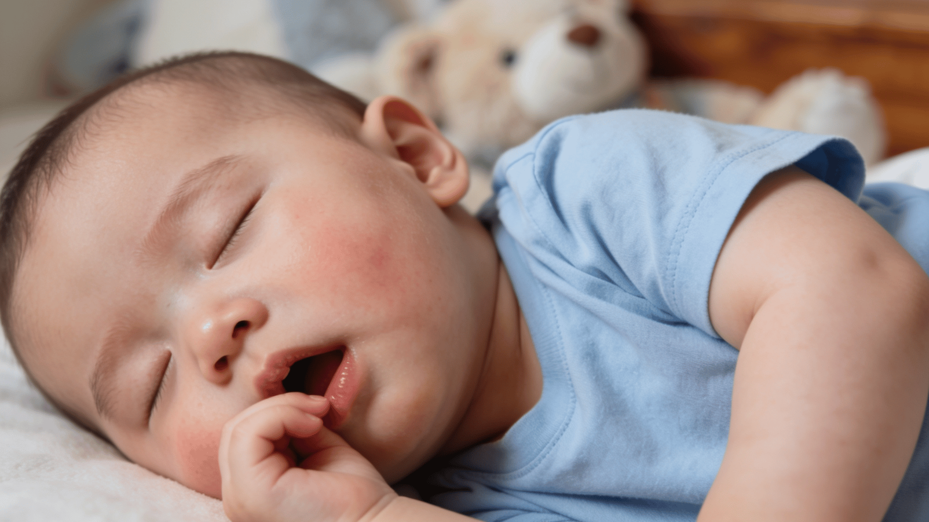 sleeping baby in a blue outfit resting on a bed with rosy cheeks and a teddy bear softly blurred in the background