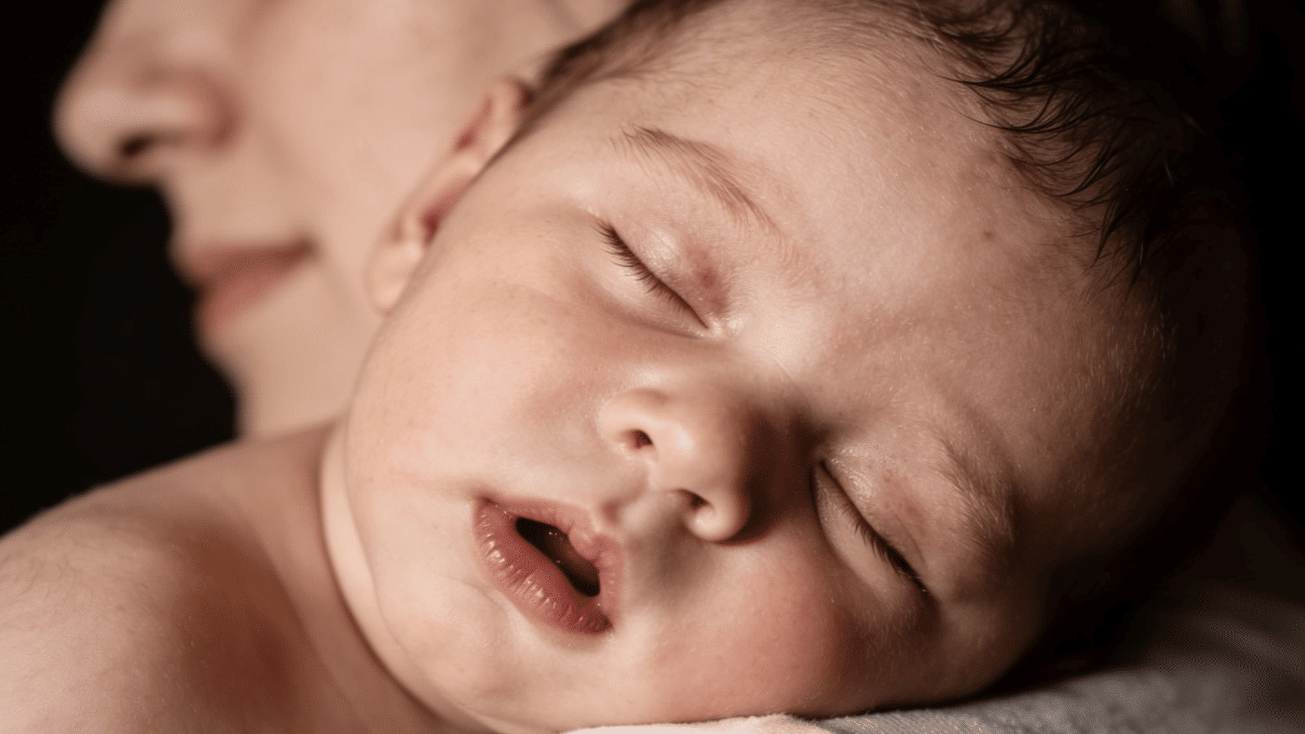 sleeping newborn baby resting on a caregiver’s shoulder with closed eyes and a peaceful expression in soft warm light