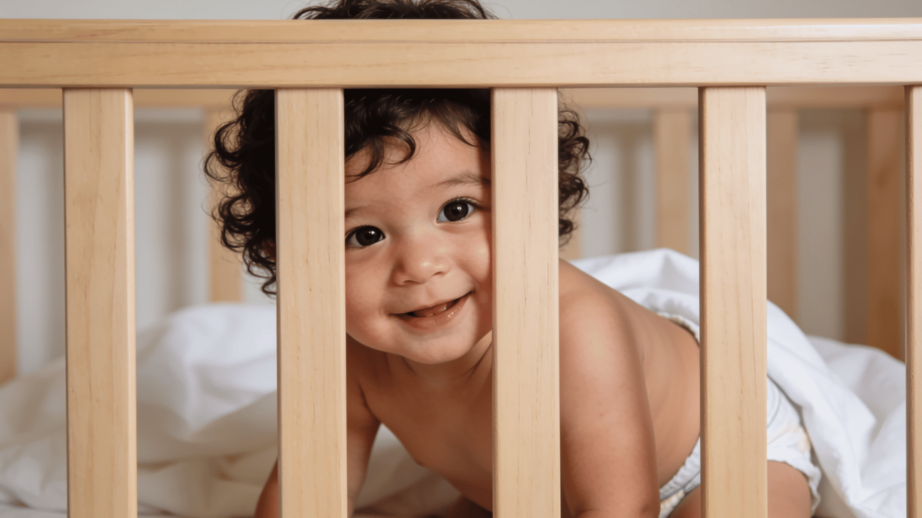 smiling baby standing inside a wooden crib wearing a diaper and looking through the crib rails with curiosity