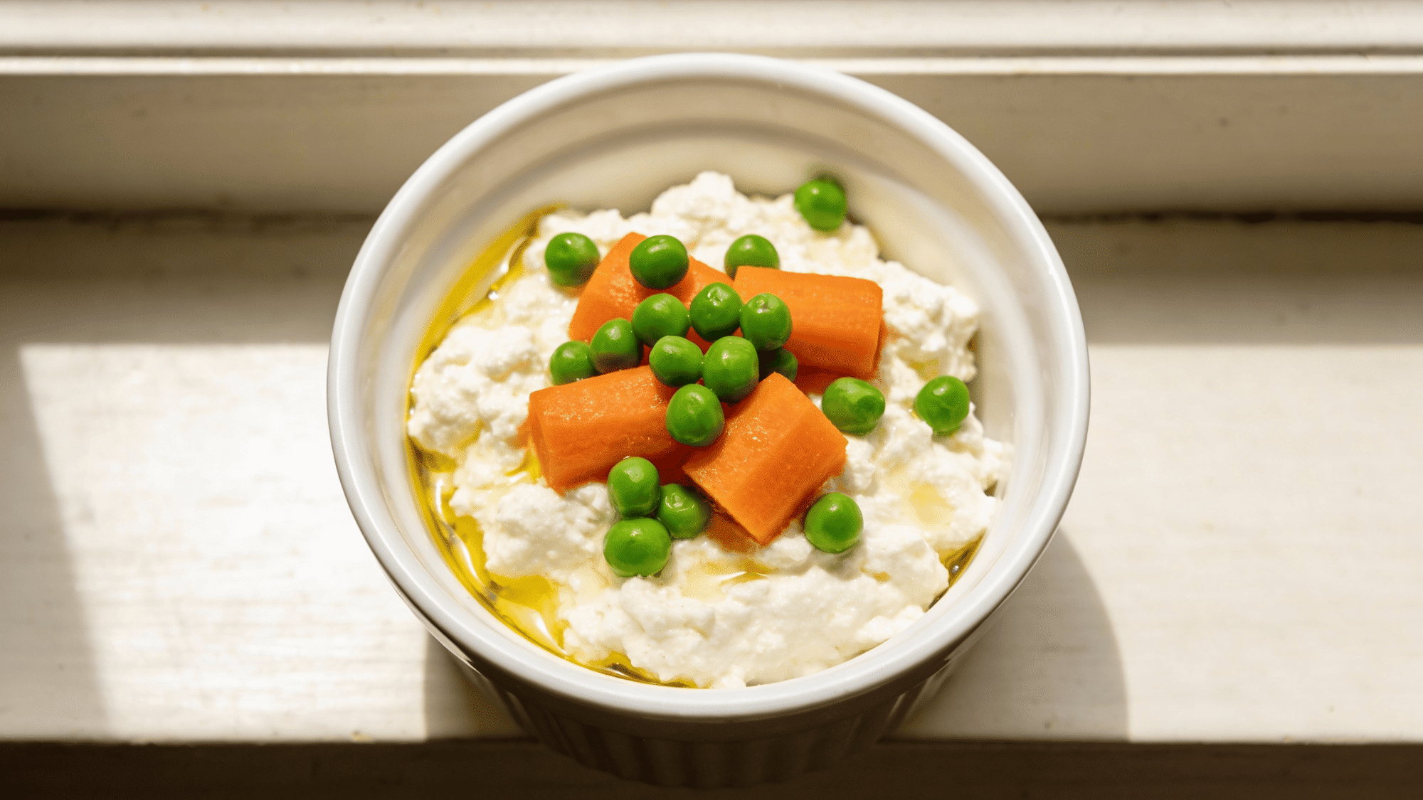 white bowl of cottage cheese topped with steamed carrots, green peas, and a drizzle of oil