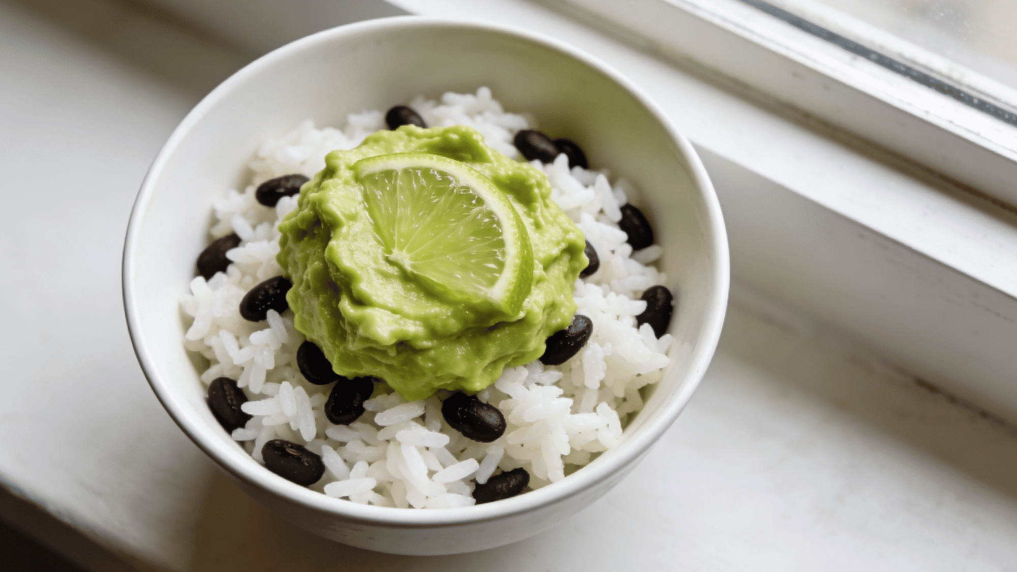 white bowl of white rice and black beans topped with smooth mashed green avocado