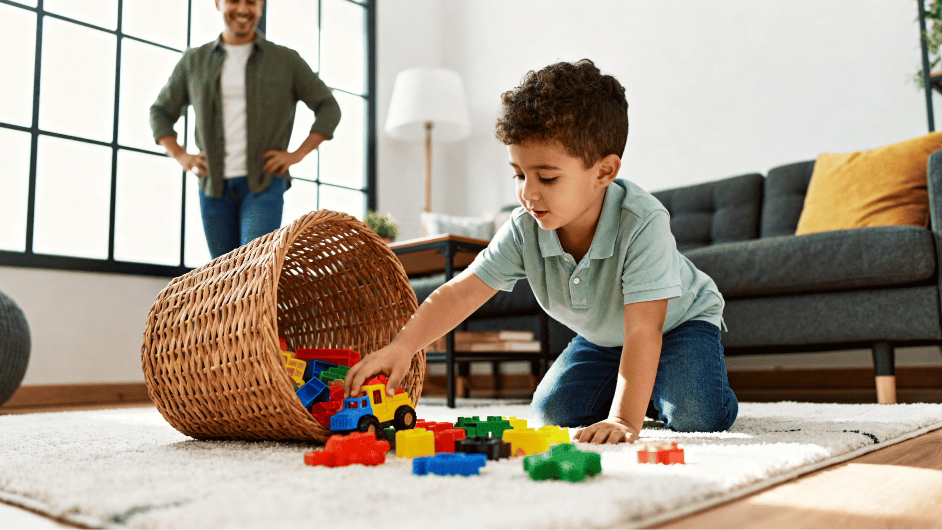 young child cleaning up toys independently while supportive parent watches proudly in a bright family room.