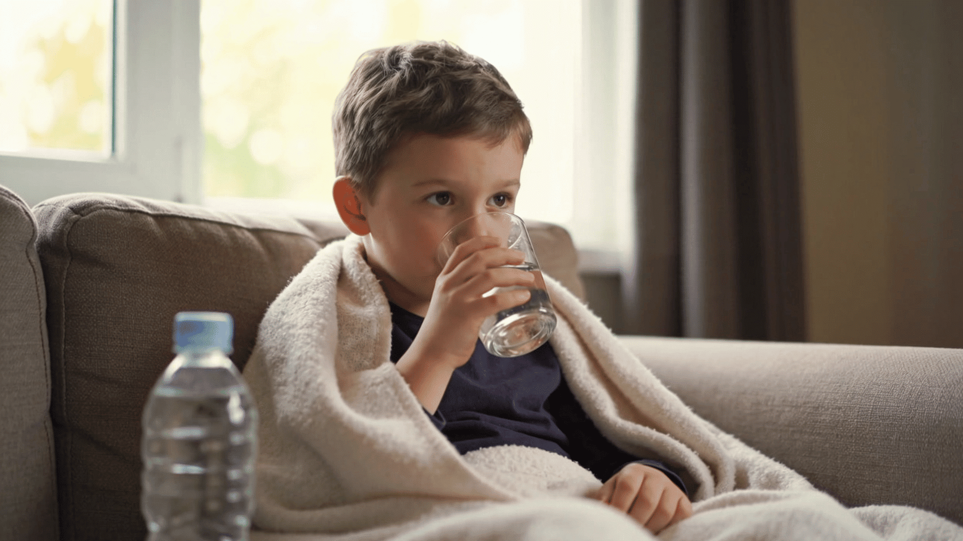 child drinking water while resting under a blanket at home during illness recovery and staying hydrated