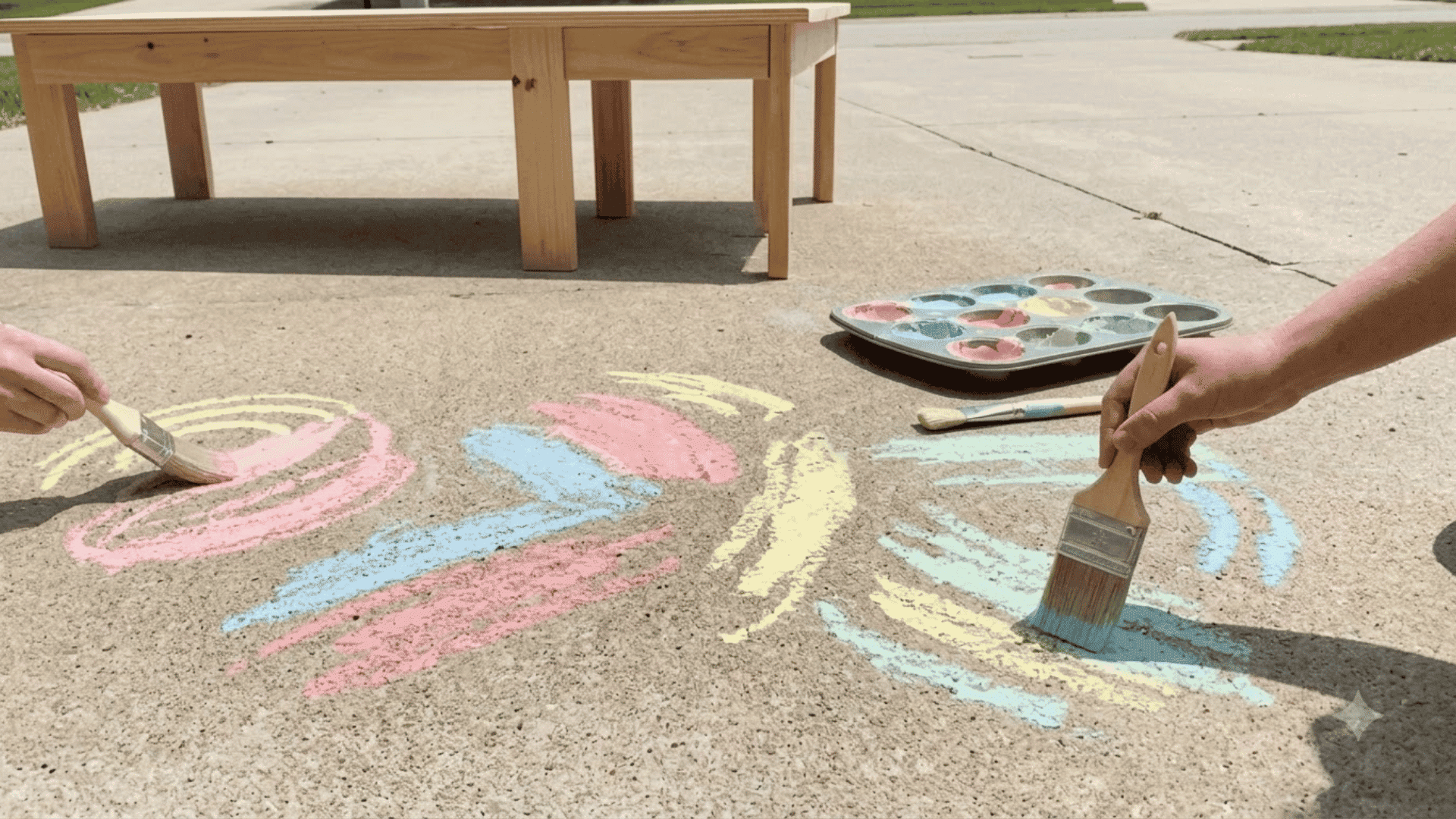 children paint colorful pastel shapes on a sunny concrete sidewalk using brushes and a tray of washable paints