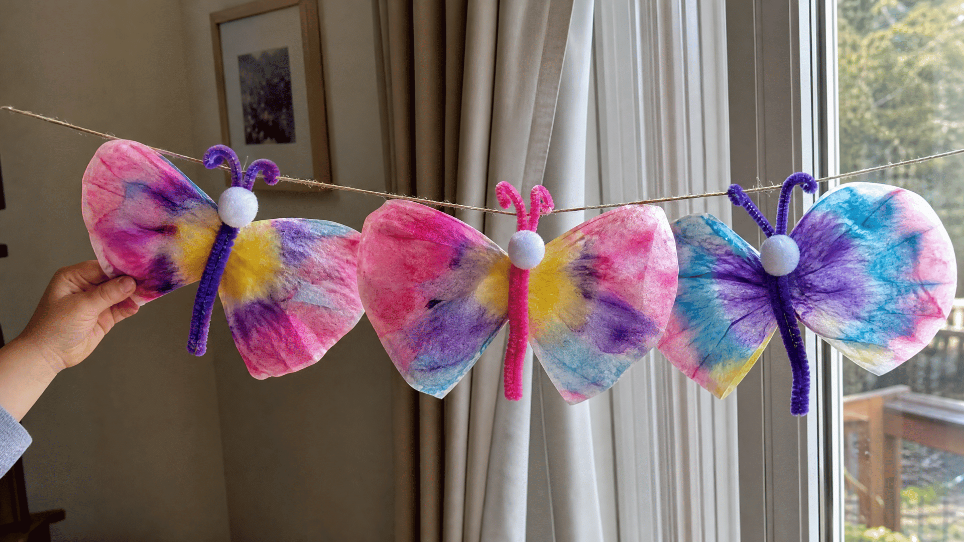 colorful coffee filter butterflies with pipe cleaner antennae hanging on a string near a window