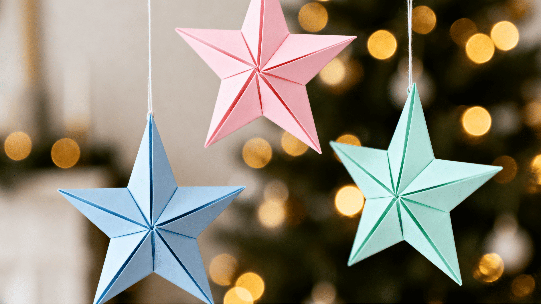 colorful handmade origami star ornaments in pastel pink blue and green hanging on white strings soft bokeh christmas lights in blurred background