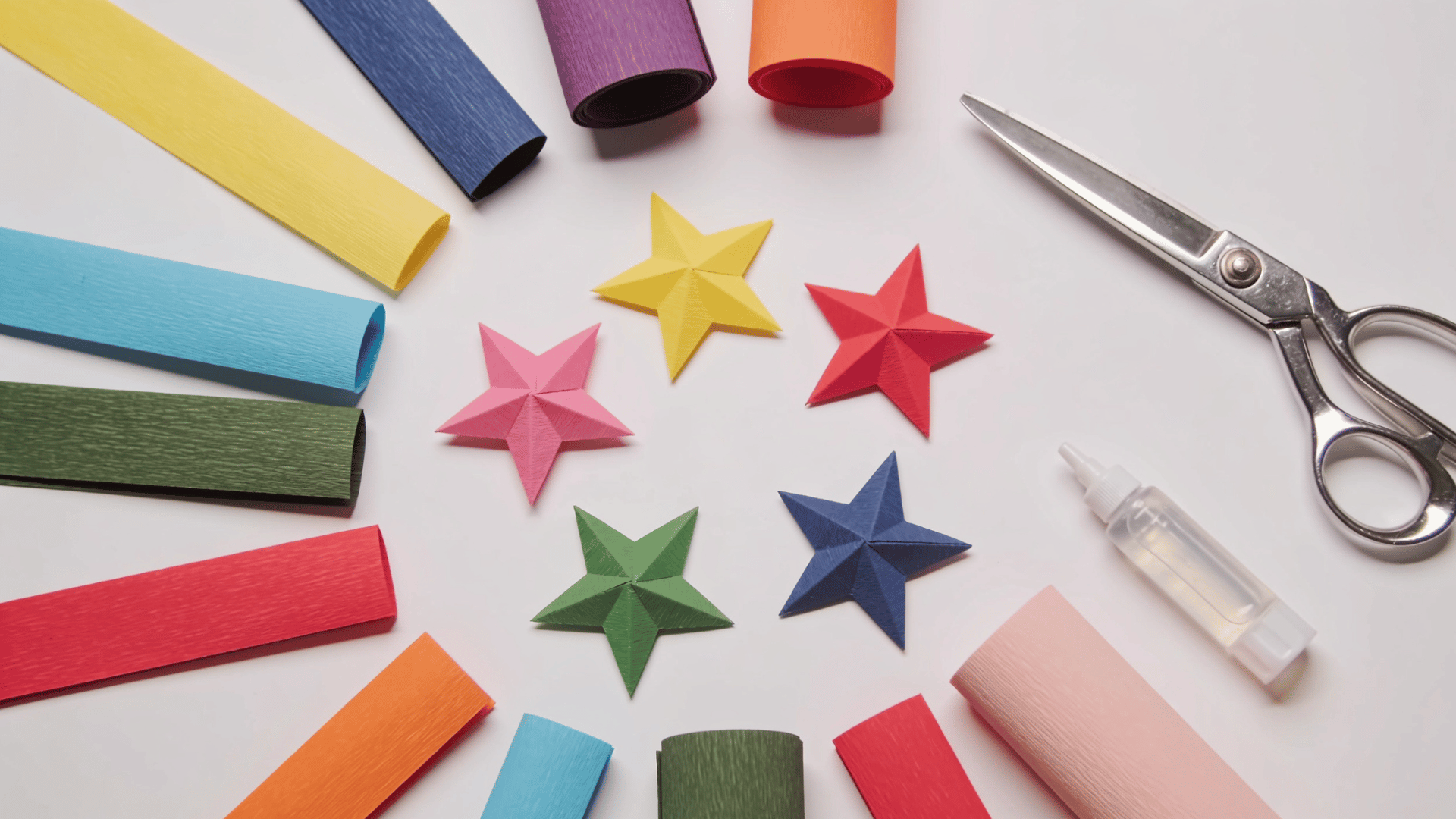 colorful handmade paper stars surrounded by craft paper strips, scissors, and glue on a clean white workspace table