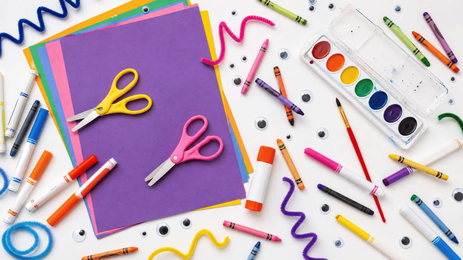 colorful kids craft supplies scattered on white background including scissors, crayons, markers and googly eyes