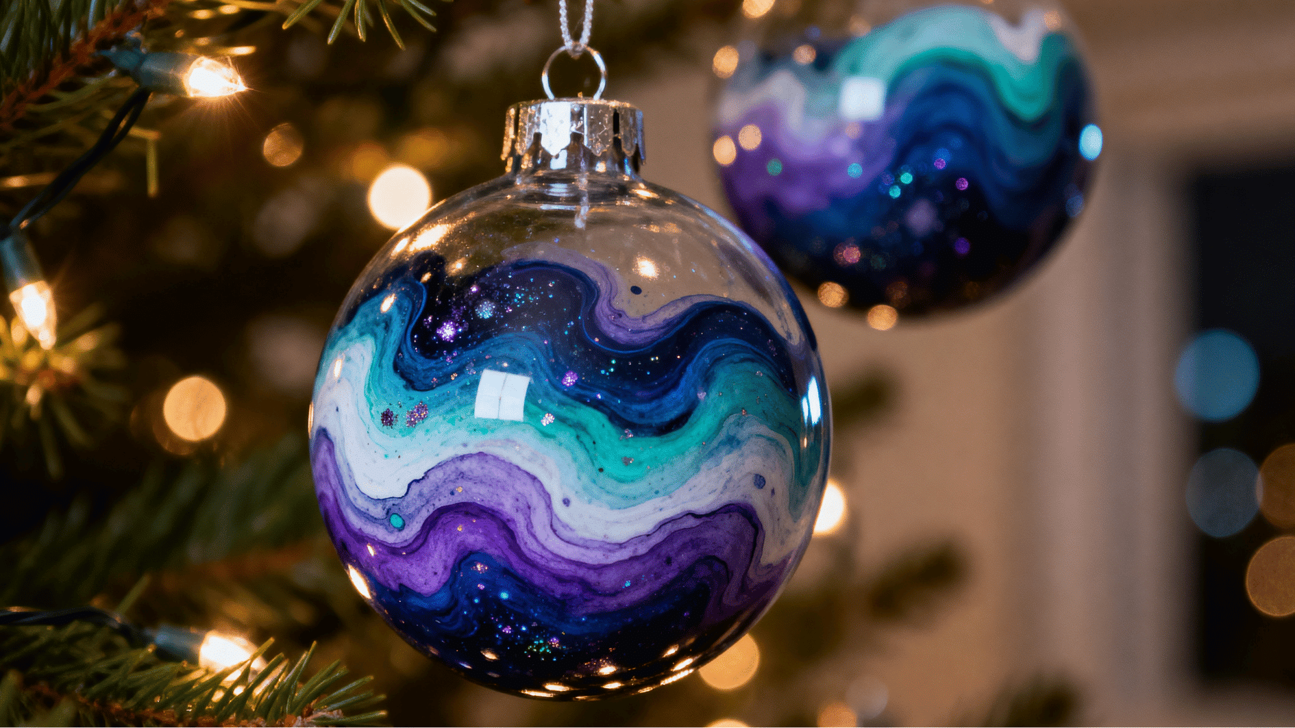 glass christmas bauble ornament with swirling blue purple teal galaxy paint design hanging on tree with warm lights background