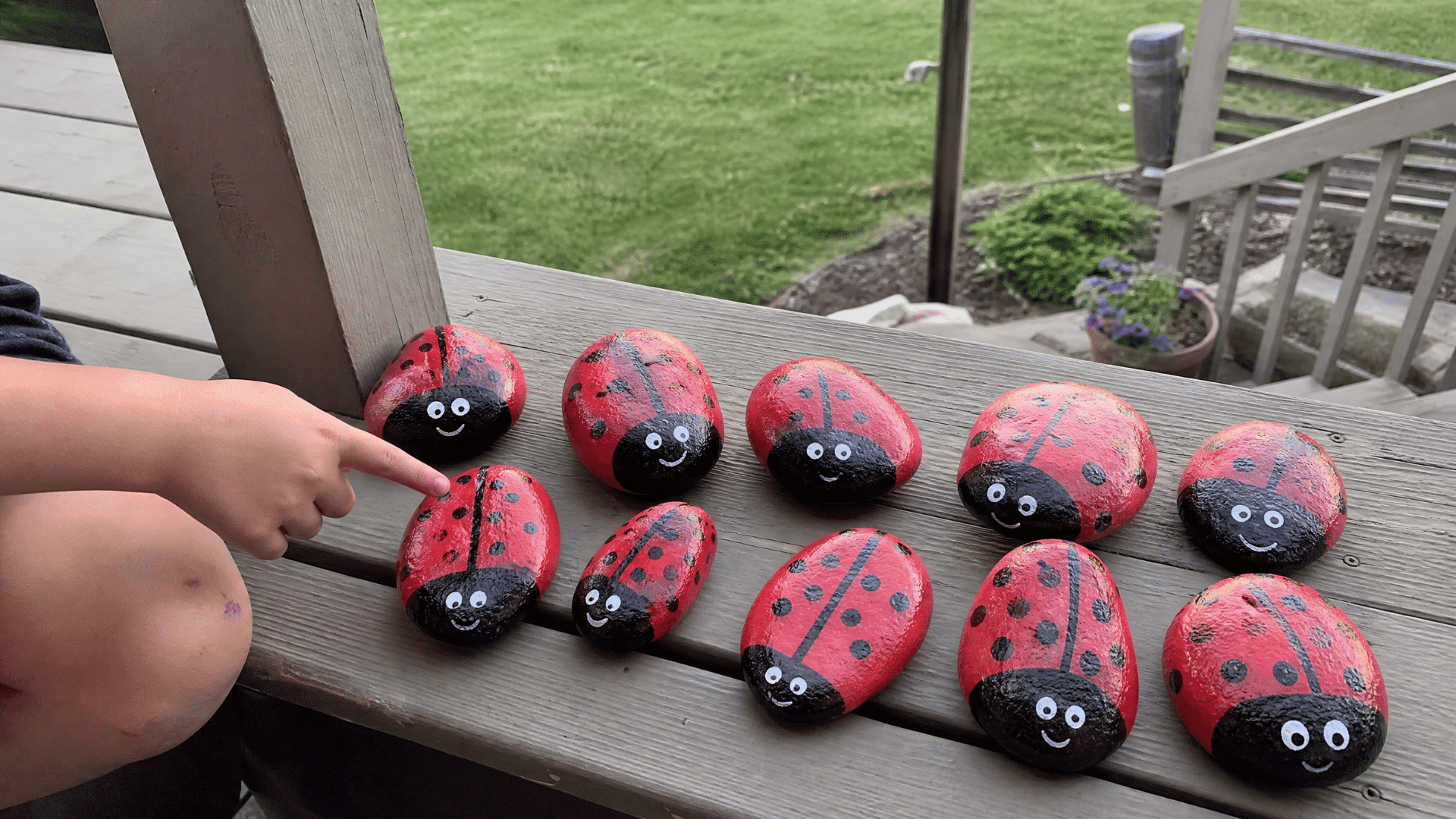 kids painted ladybug rocks arranged on wooden deck railing with american backyard and grass in background