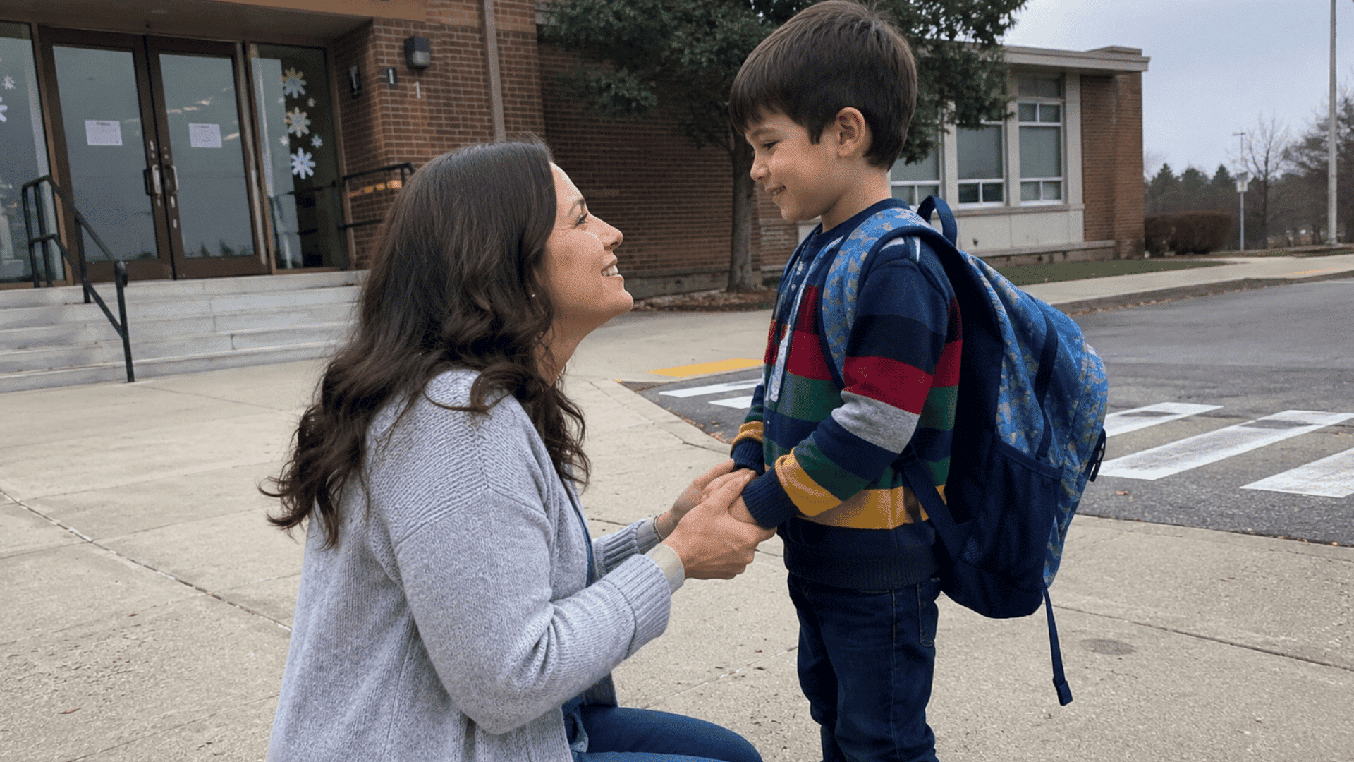 mom kneeling and holding her son's hands outside an elementary school on his first day of daycare drop-off