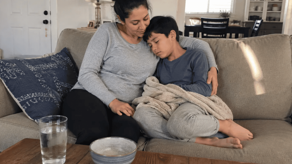 mother comforting her tired, unwell child resting on the couch with a blanket, water and bowl nearby