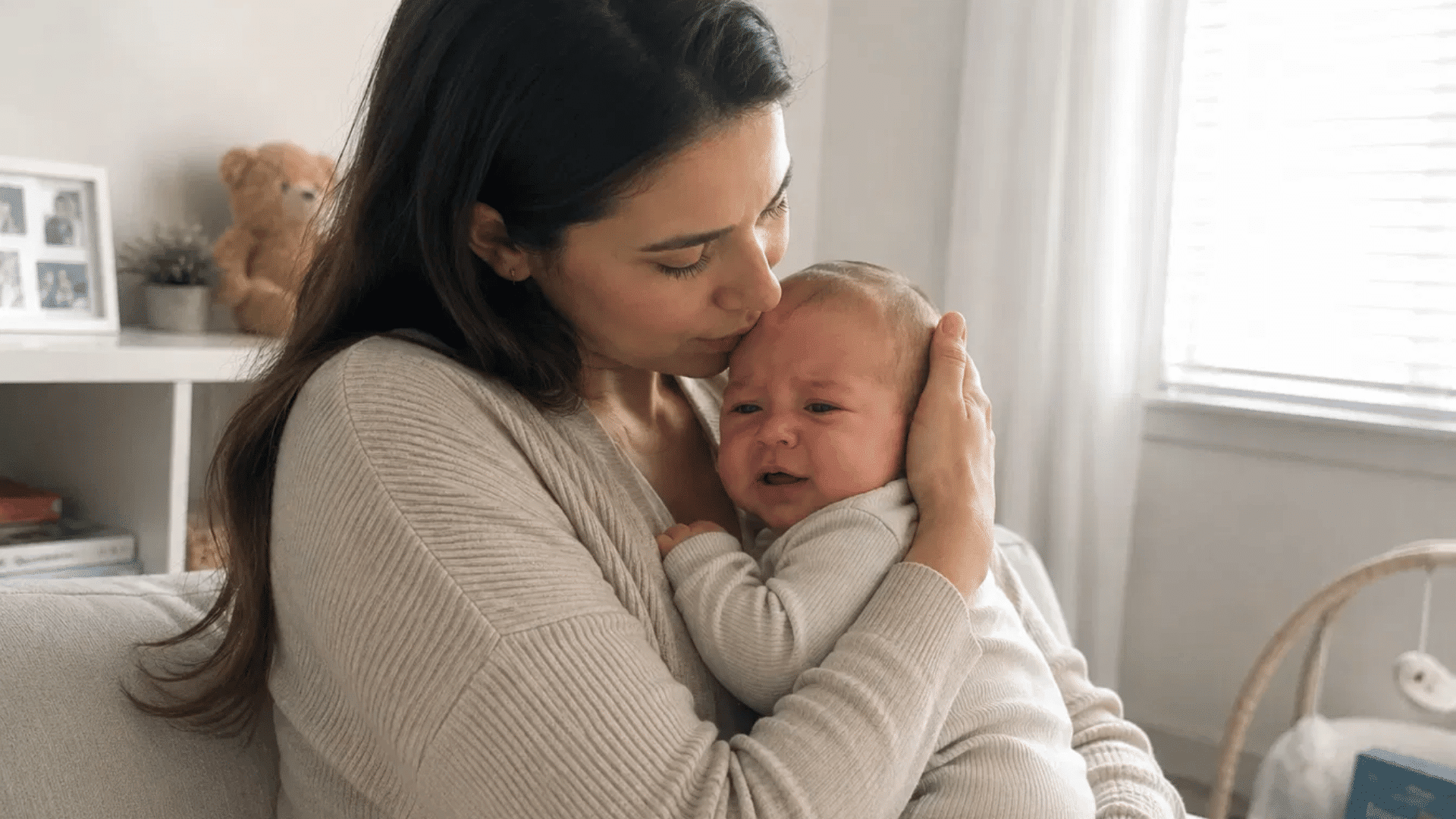 mother gently kissing and soothing a fussy newborn on a couch in a bright cozy living room