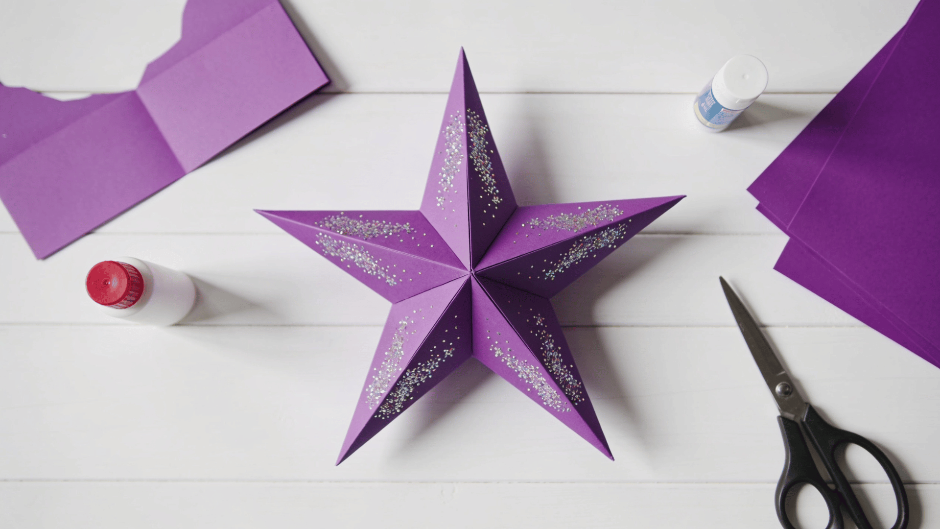 purple five-point paper star decorated with glitter, surrounded by craft paper, glue, and scissors on a white table