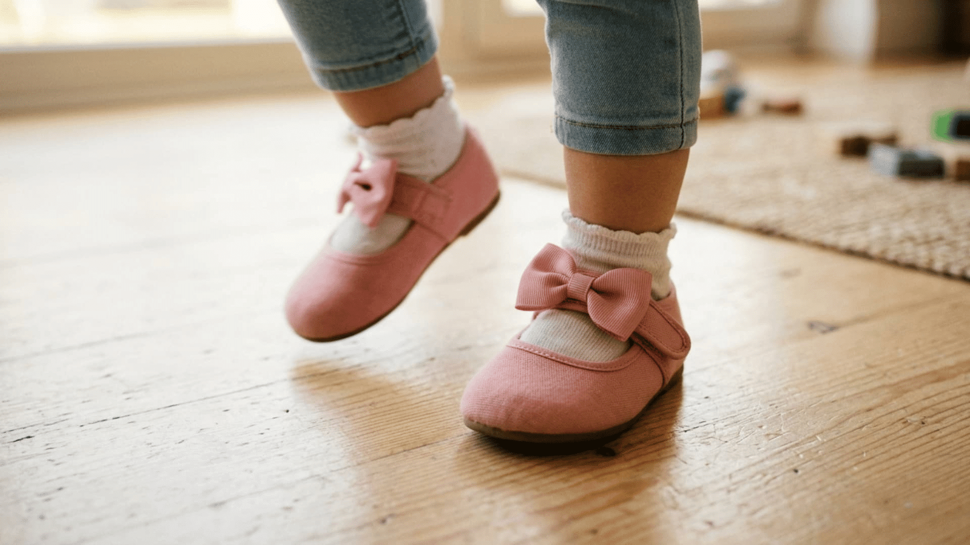 toddler girl walking indoors in pink bow shoes white socks and rolled blue jeans on a wooden floor
