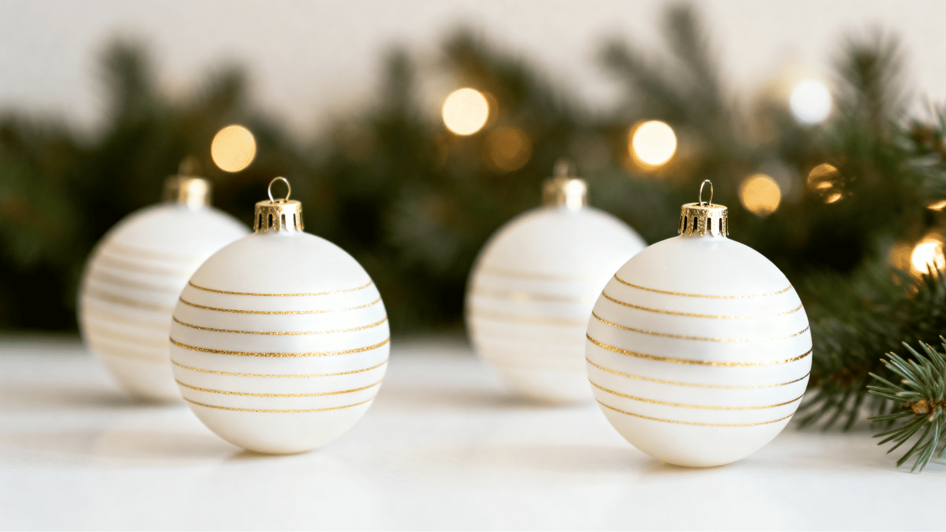 white christmas baubles with thin gold stripes arranged on soft surface with evergreen branches and warm festive lights bokeh