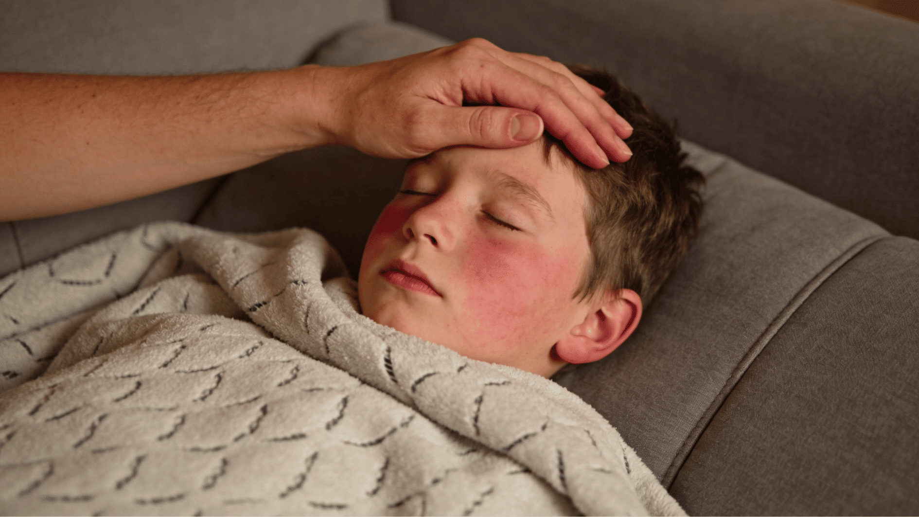 young child lying on grey sofa with fever with parent hand on his head in warm indoor light