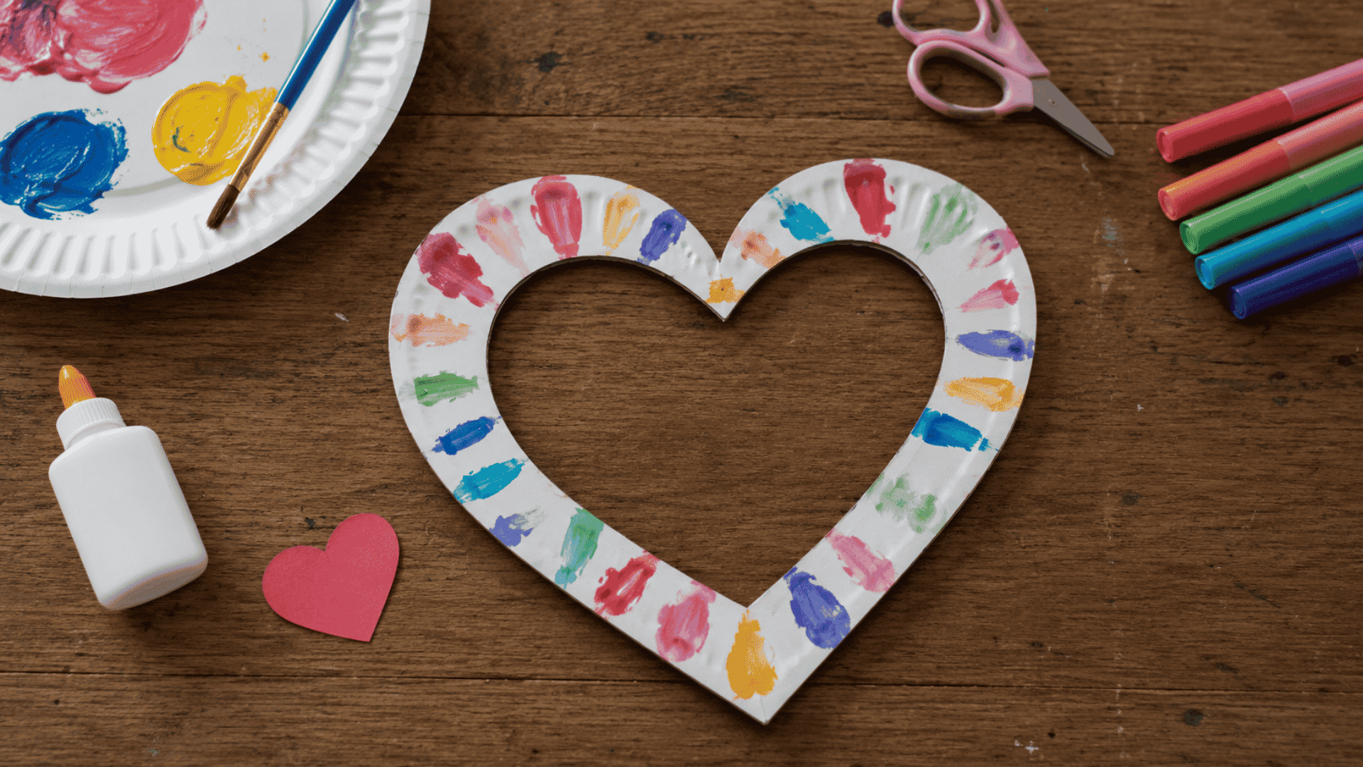 a handpainted heart shaped photo frame with colorful brush marks and craft supplies arranged on a wooden table